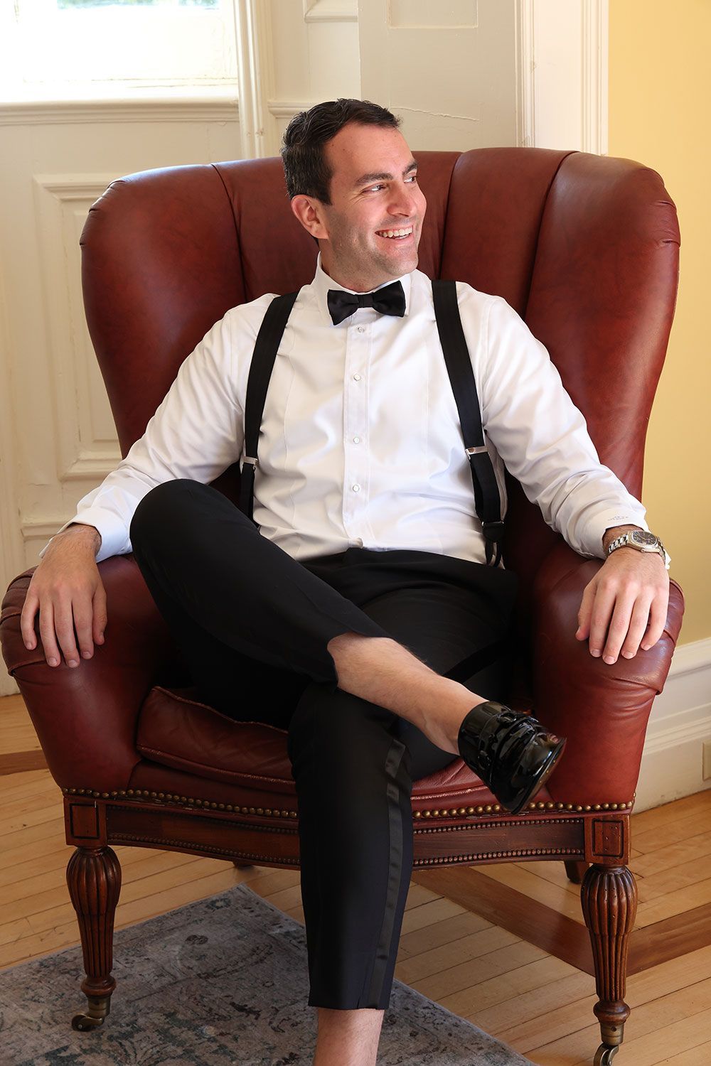 Man in formal attire, seated in a red leather armchair, looking to the side and smiling.