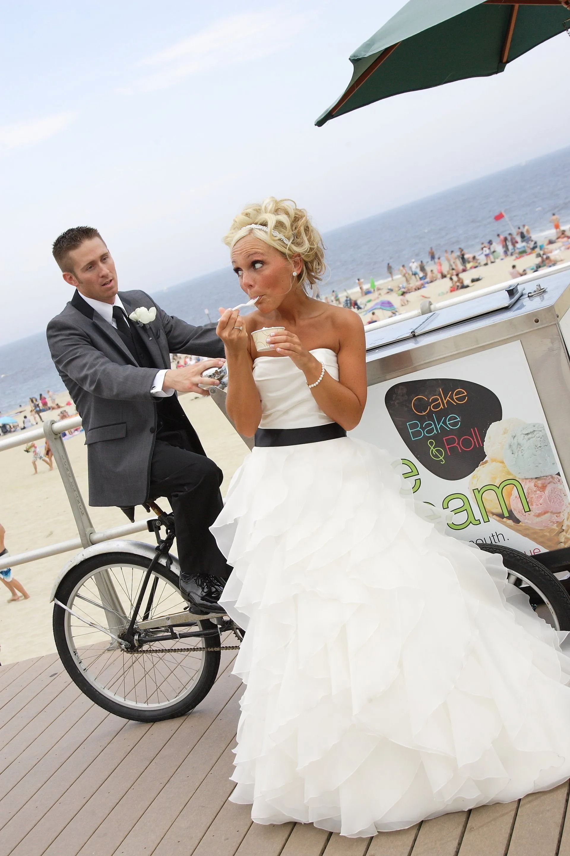 Groom riding a bike, bride eating ice cream on a boardwalk near a beach.