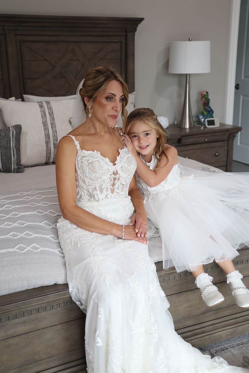 Woman in wedding dress sits on bed with flower girl in white dress.