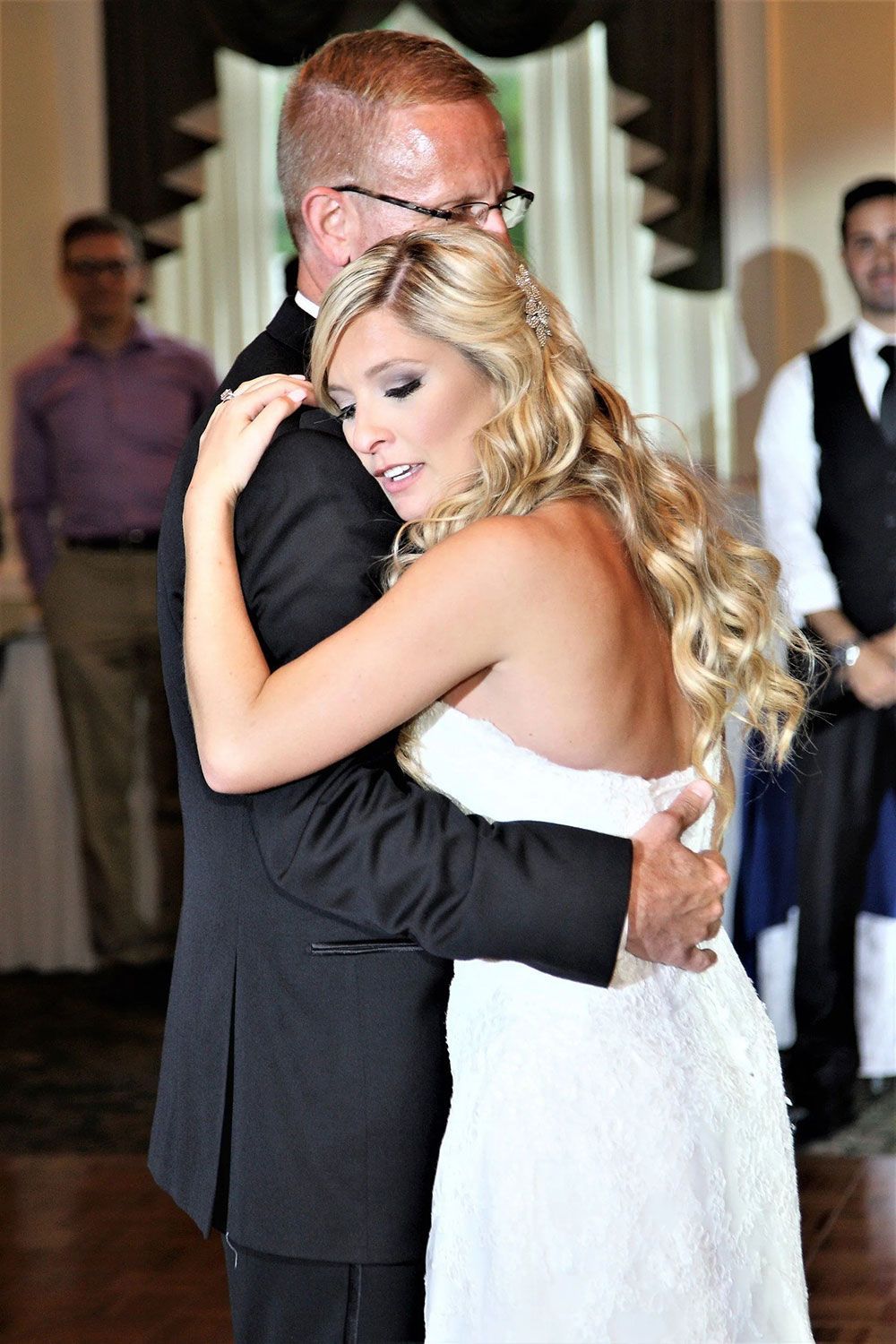 Bride in white dress hugs a man in a black suit, dancing at a wedding reception.