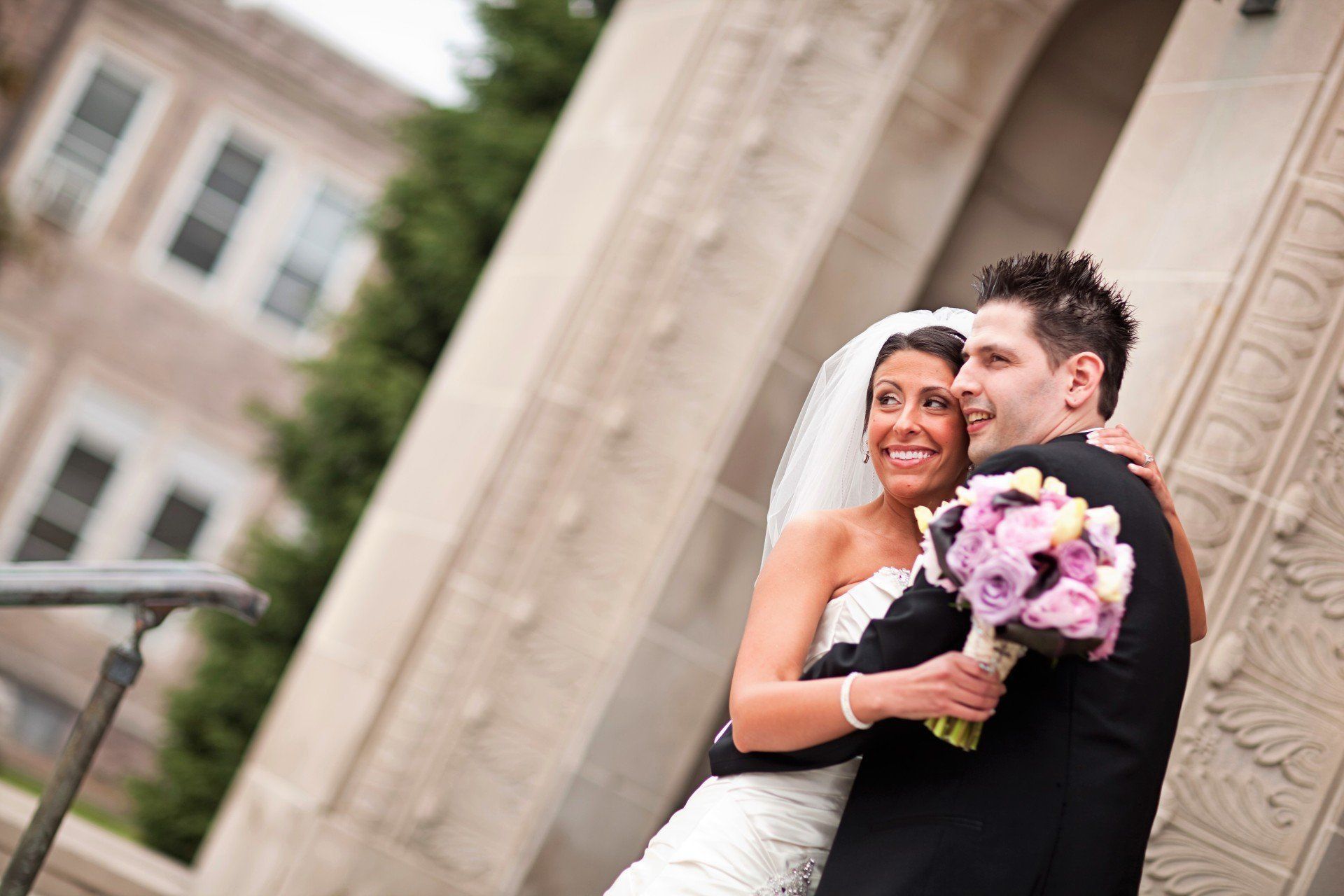 Newlyweds embracing, bride holding purple bouquet, standing outside stone building, happy expressions.