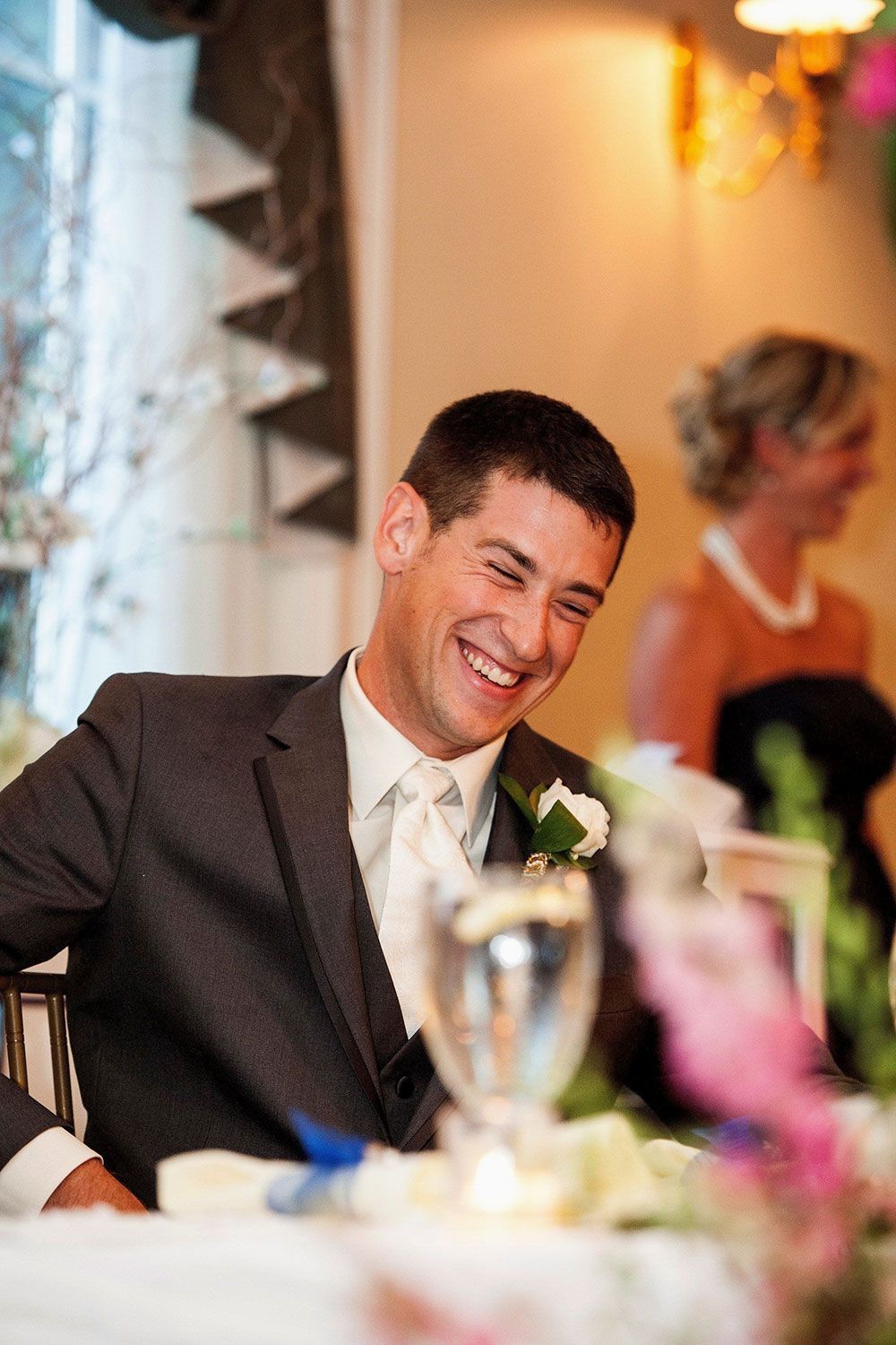Man in a suit smiles widely at a table during an event. A woman is in the background.