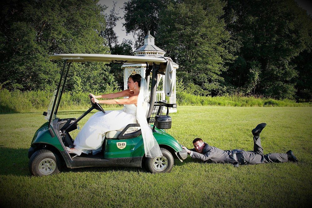 Bride driving a golf cart, pulling groom on ground; outdoor wedding.