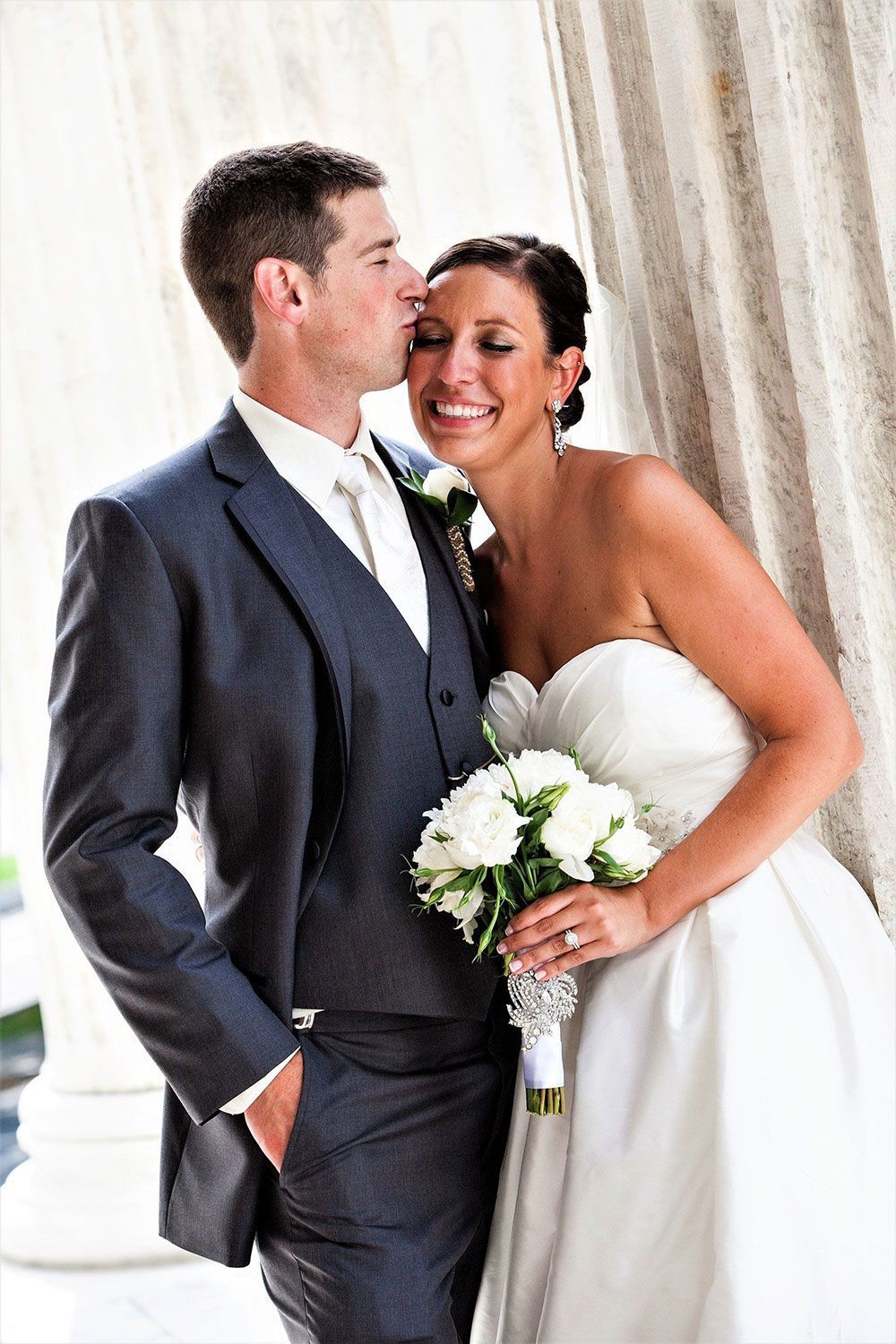 Groom kissing bride's forehead. Bride in strapless dress holds bouquet, both smiling near a white column.