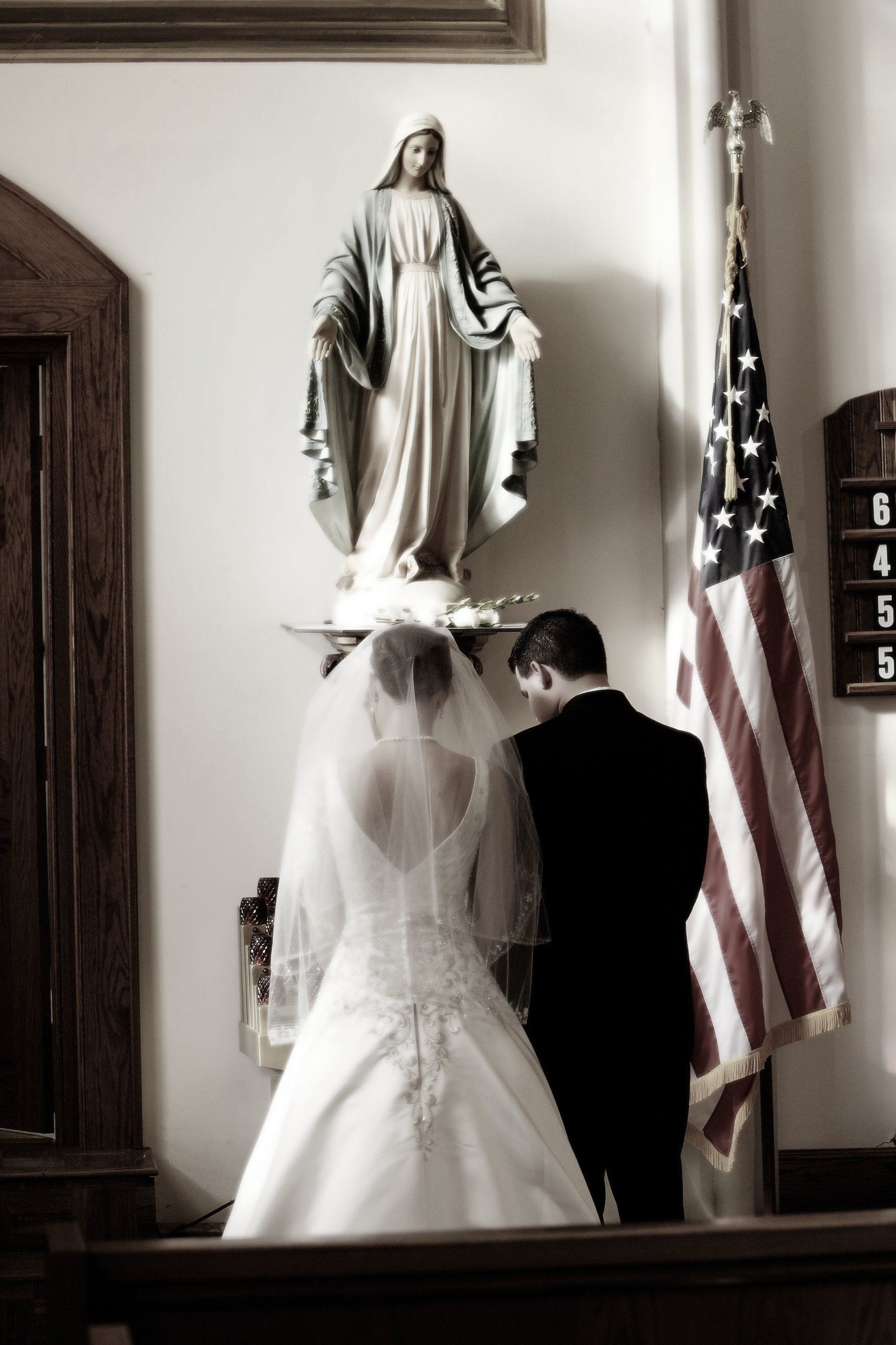 Couple in wedding attire praying before a statue of the Virgin Mary in a church; American flag nearby.
