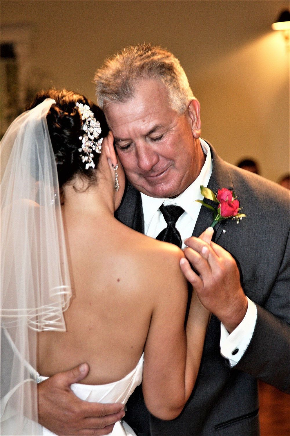 Bride and her father dance at a wedding reception; she wears a veil, he wears a suit, and they embrace.