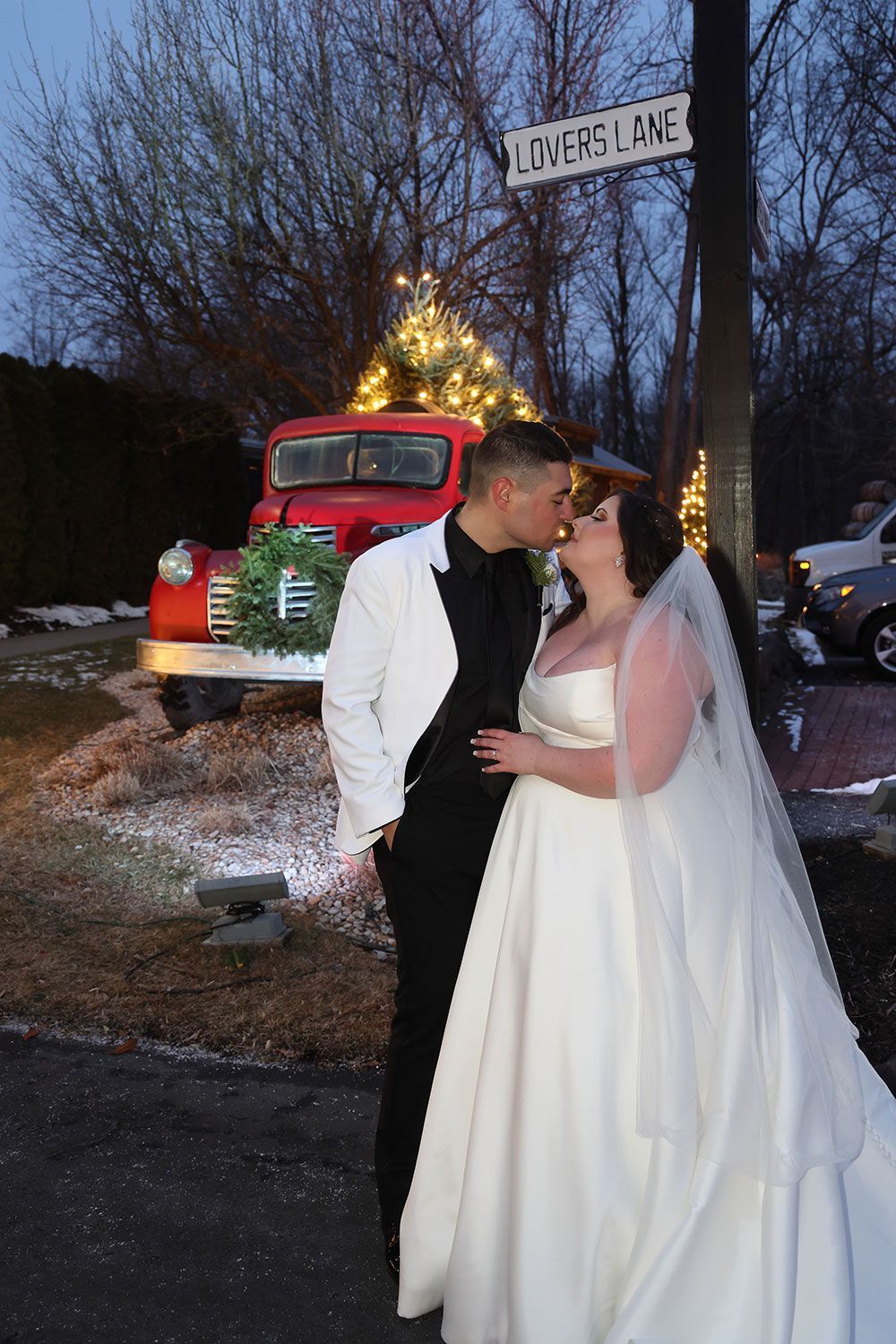 Bride and groom kissing outdoors near a red vintage truck decorated with Christmas lights and a 