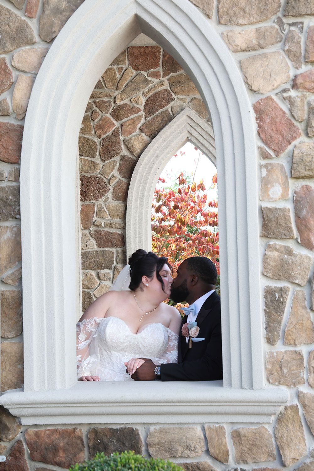 Bride and groom kissing in a stone window frame. Red leaves in background.