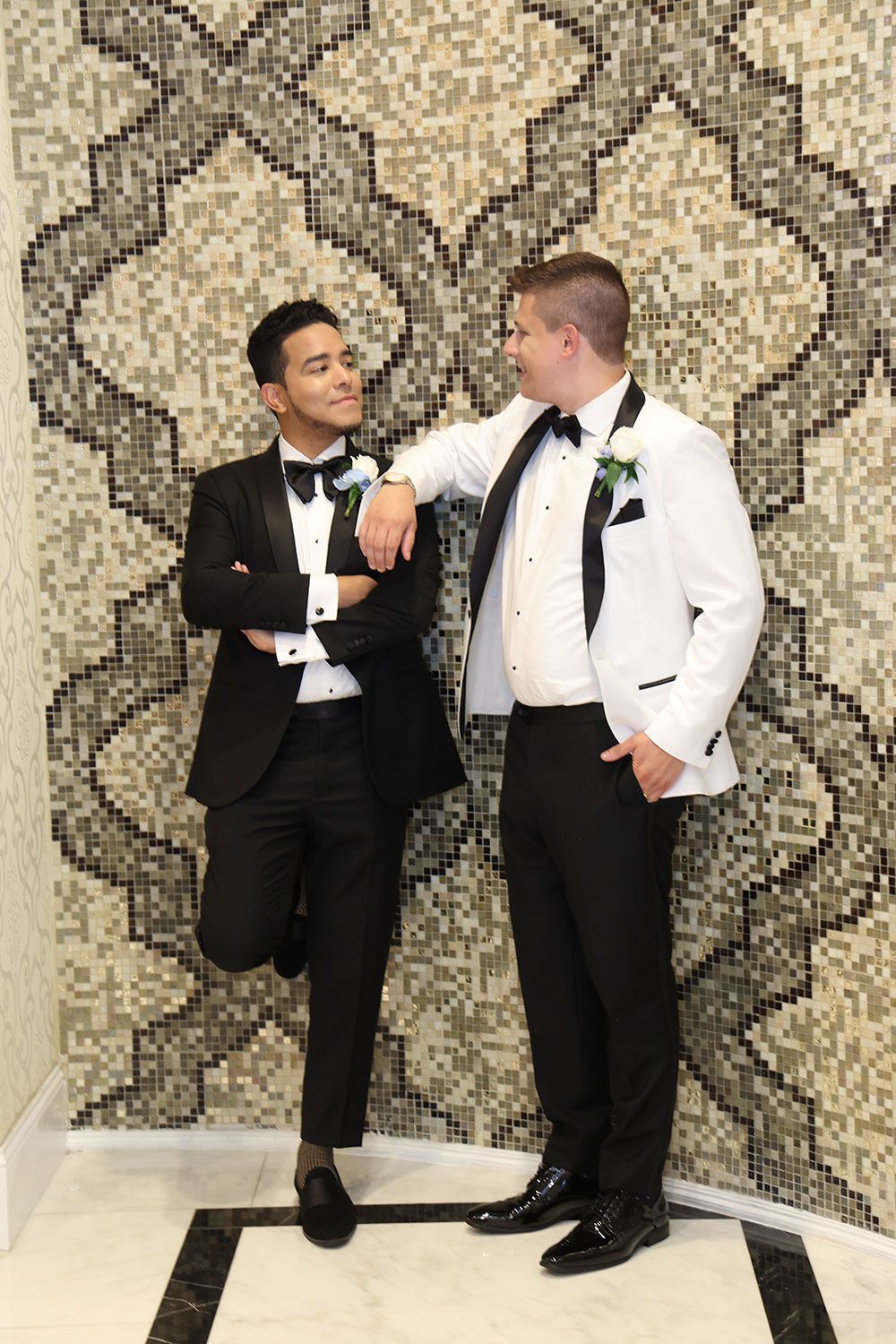 Two men in tuxedos, one in a black suit, one in a white jacket, pose by a patterned wall.