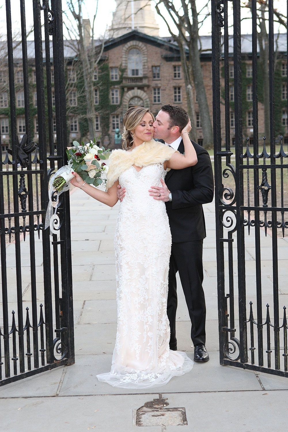 Bride and groom kissing near wrought-iron gate, holding bouquet. Building in background.