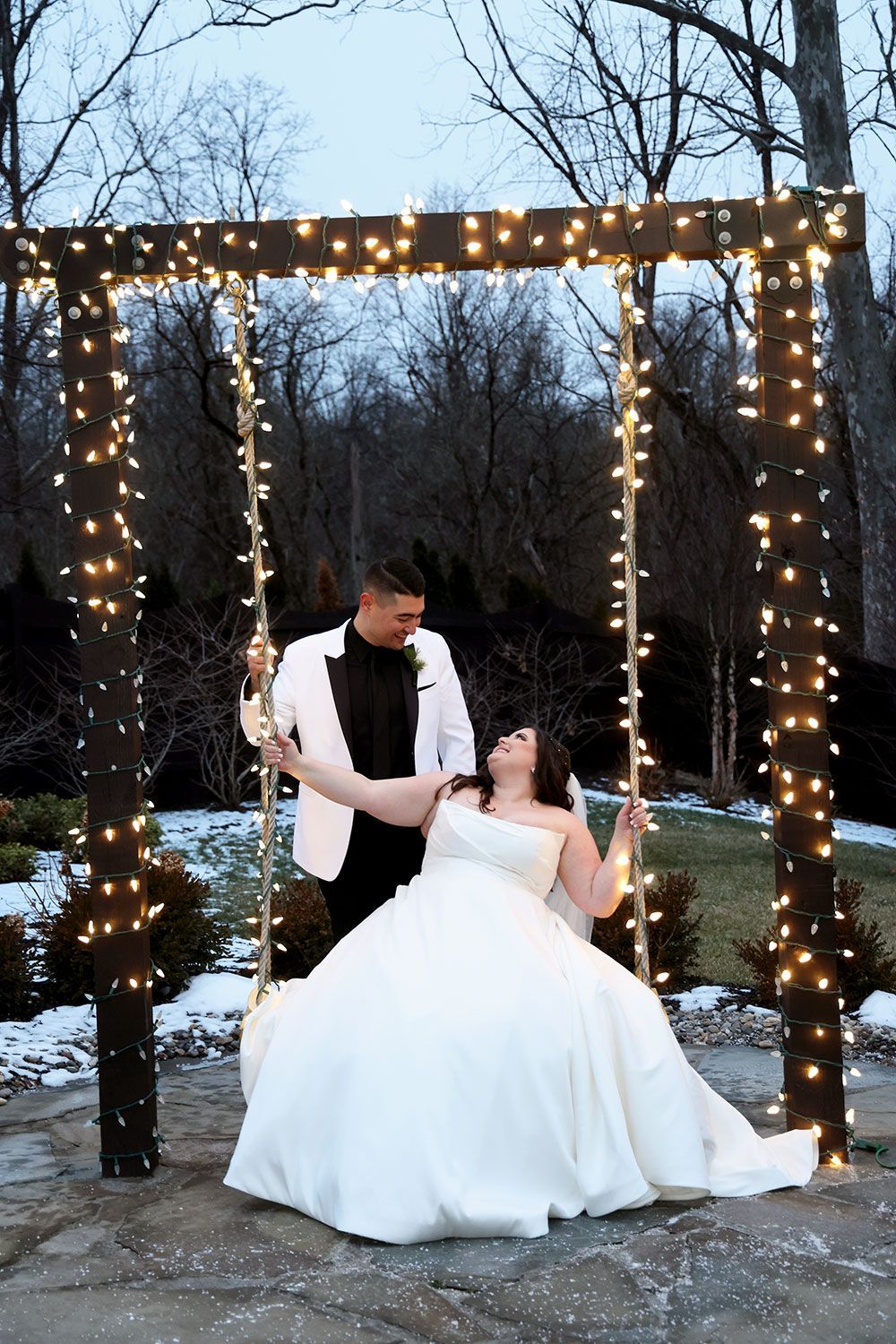 Couple in wedding attire on swing, smiling at each other, lit by string lights; outdoor setting.