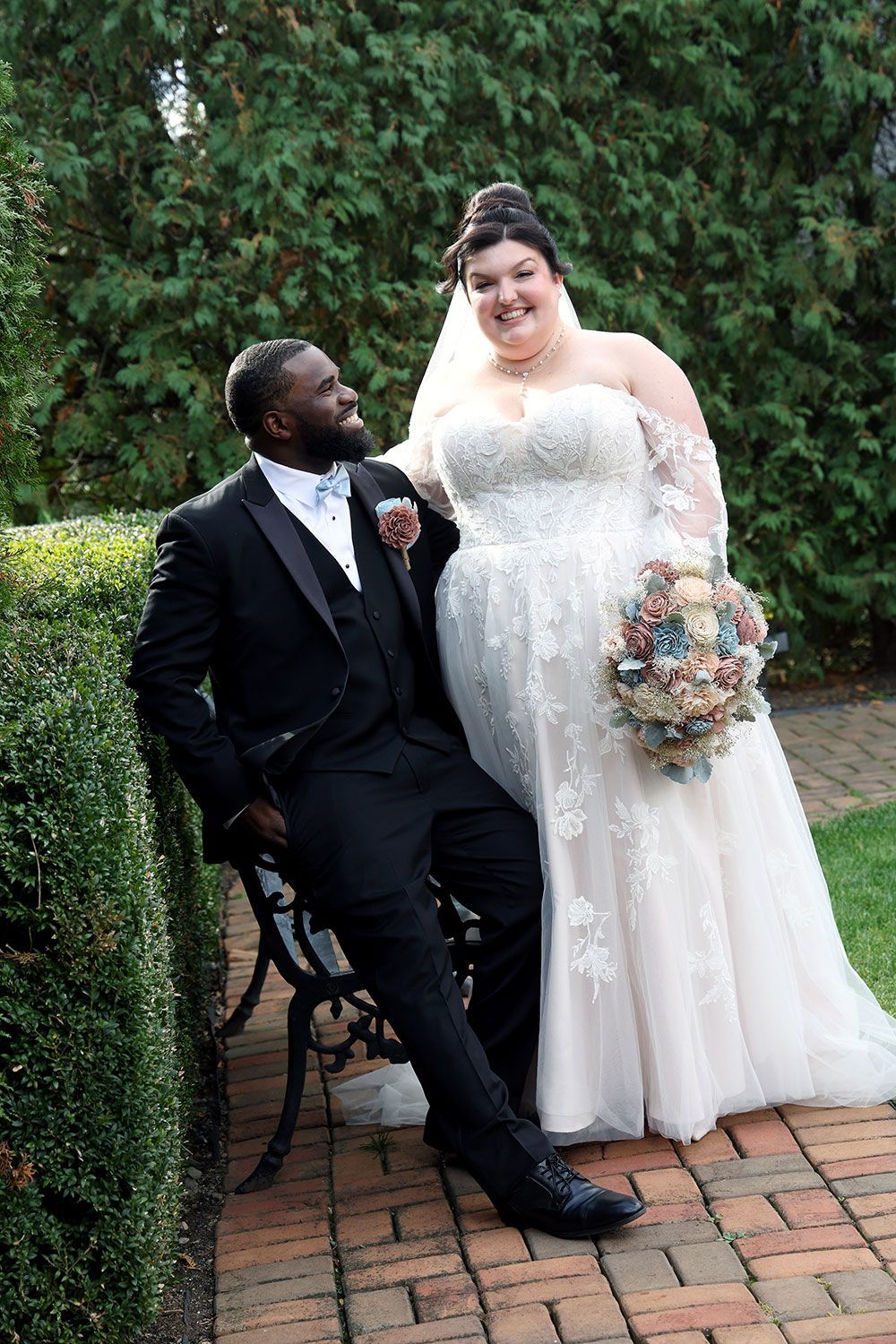 Groom in a black suit smiles up at bride in a white gown holding bouquet, both posing outdoors by greenery.