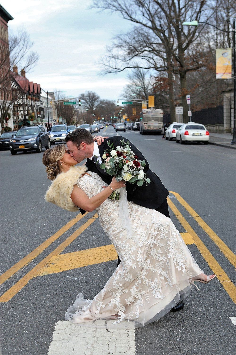 Newlyweds kiss in crosswalk, woman in white lace dress, man in suit. Busy city street.
