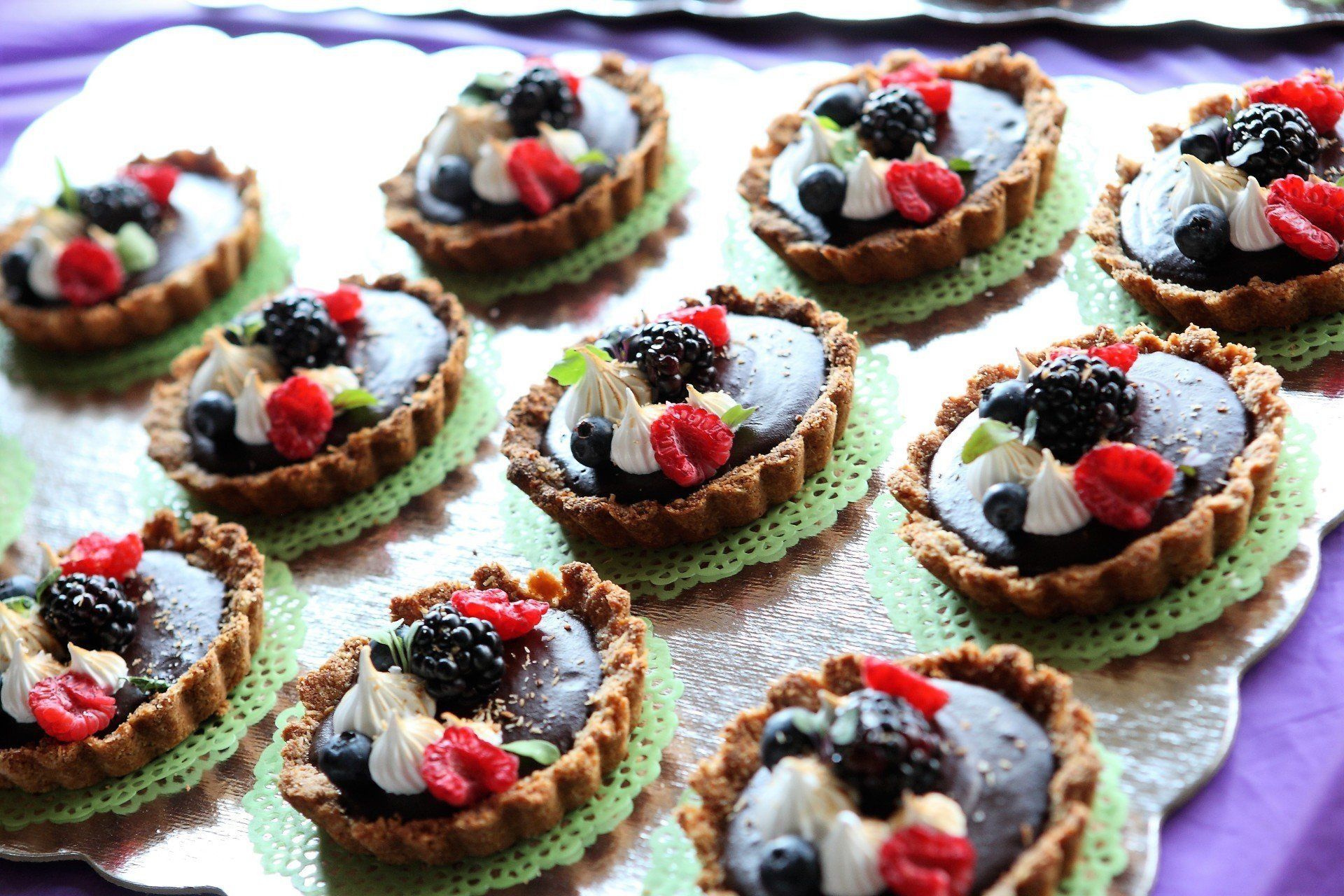 Chocolate tarts topped with berries and whipped cream on a silver platter with green doilies.