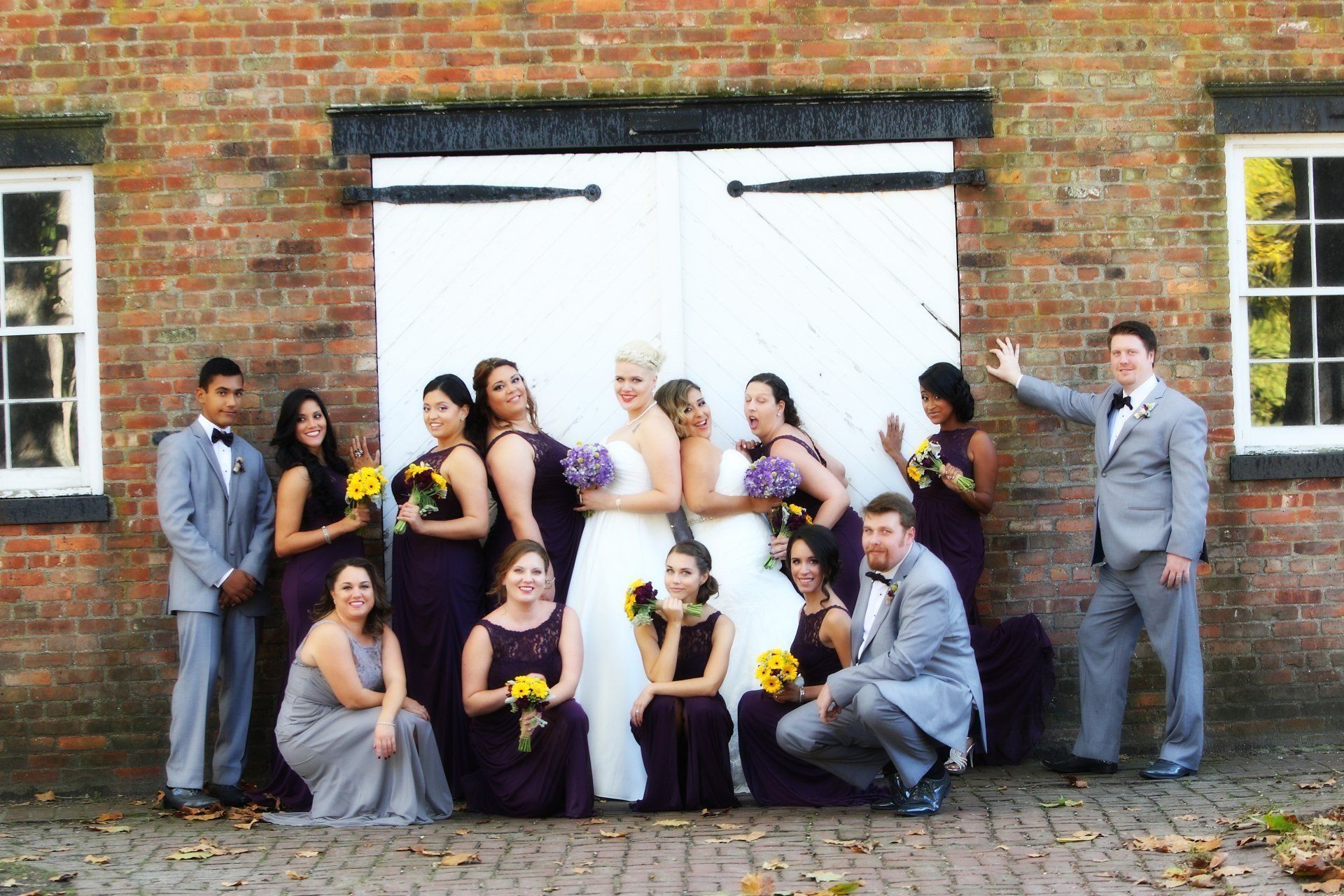 Wedding party poses in front of white barn doors. Brides wear white, bridesmaids wear purple. Groomsmen wear gray.