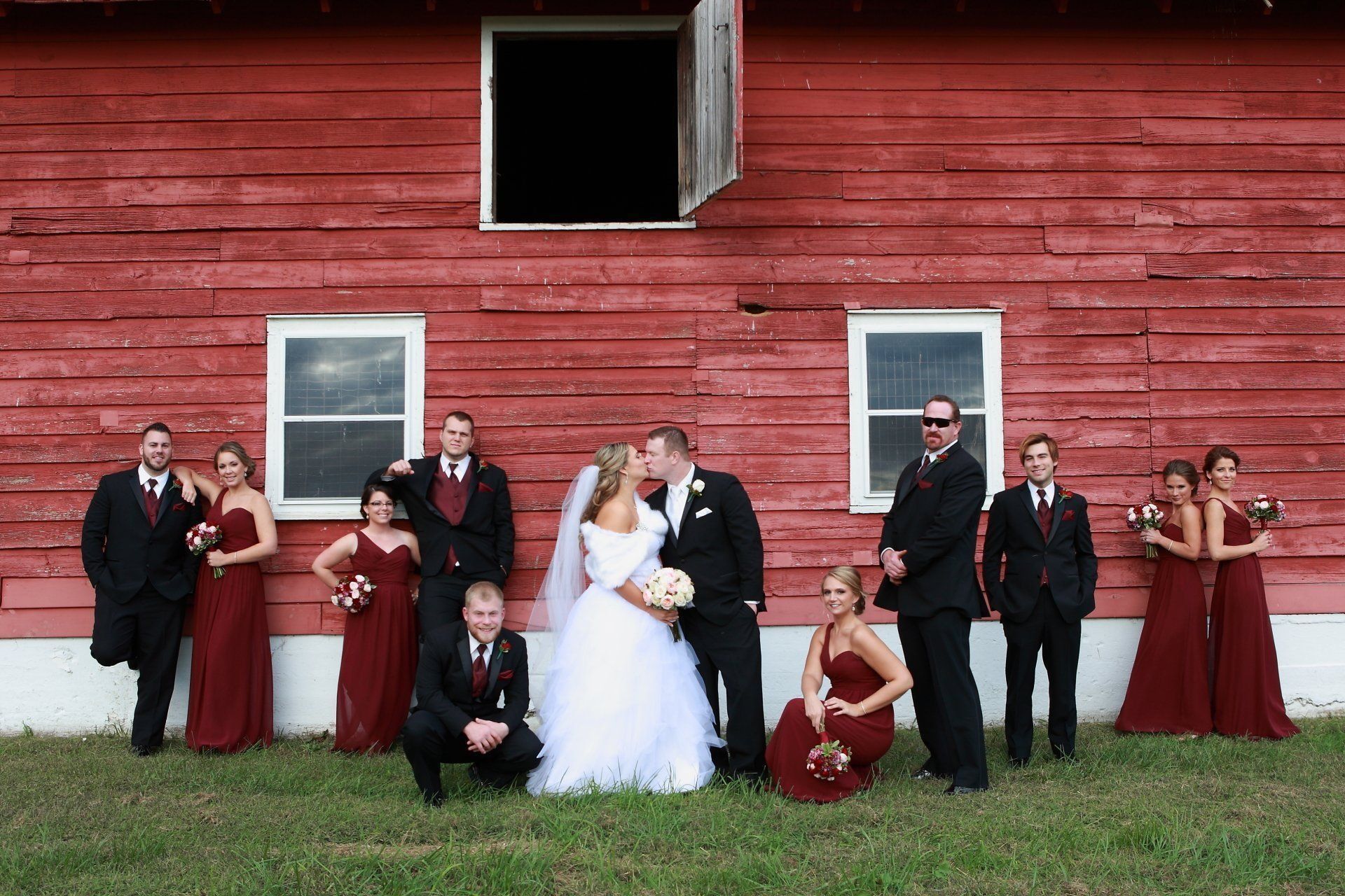 Wedding party poses in front of a red barn; bride and groom kiss, others wear black suits and maroon dresses.