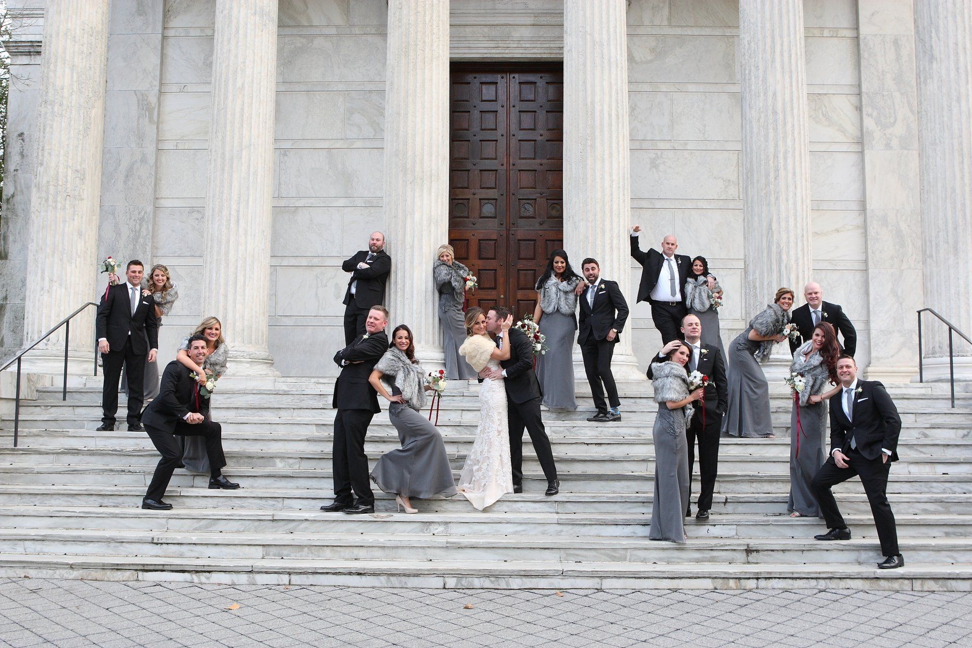 Wedding party poses on steps of a white building with columns. Guests and wedding couple act playfully.