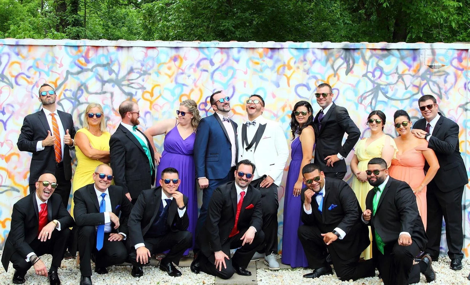 Group of people in formal attire pose in front of a colorful mural, some laughing and wearing sunglasses.