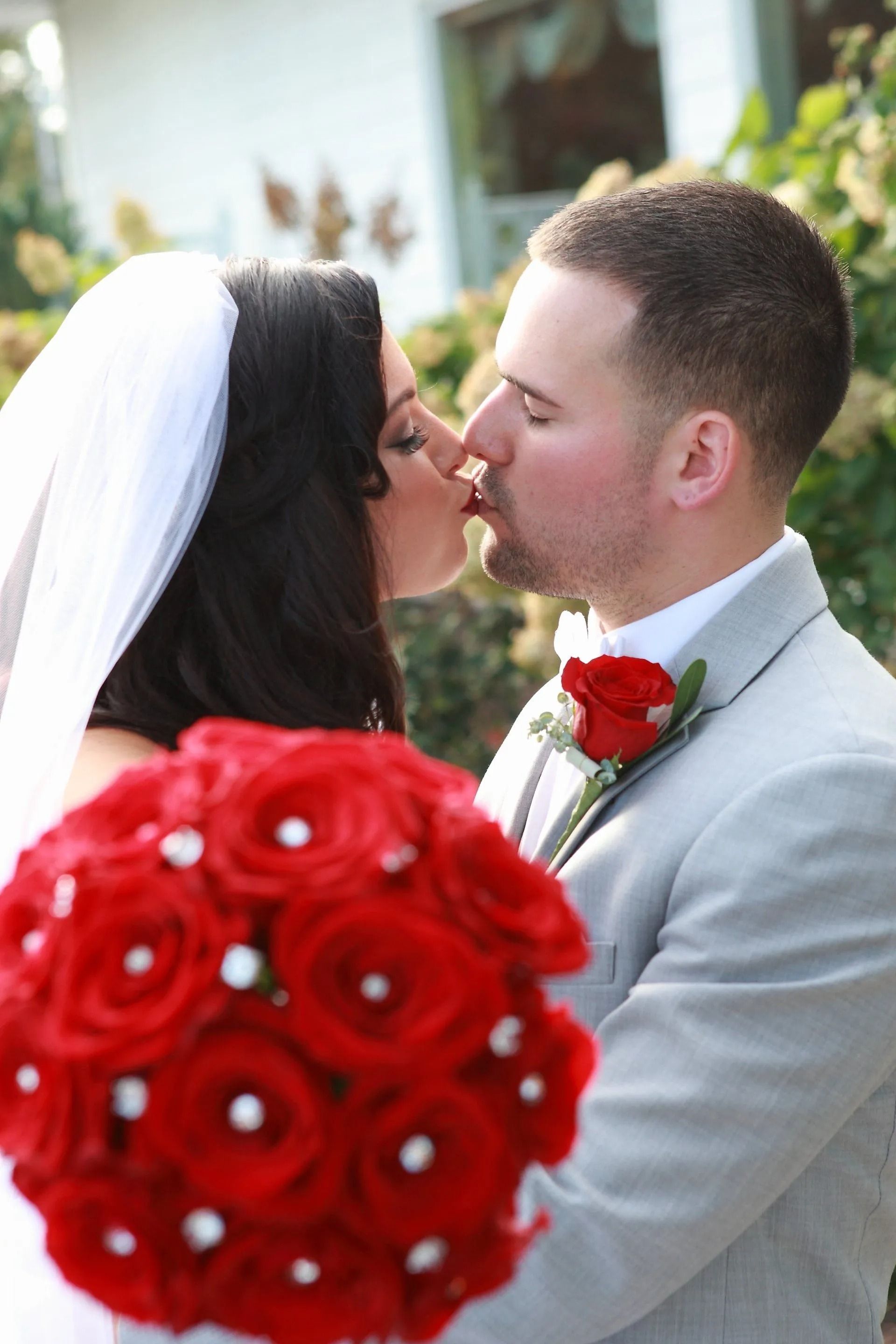Bride and groom kissing, holding red rose bouquet, outside.