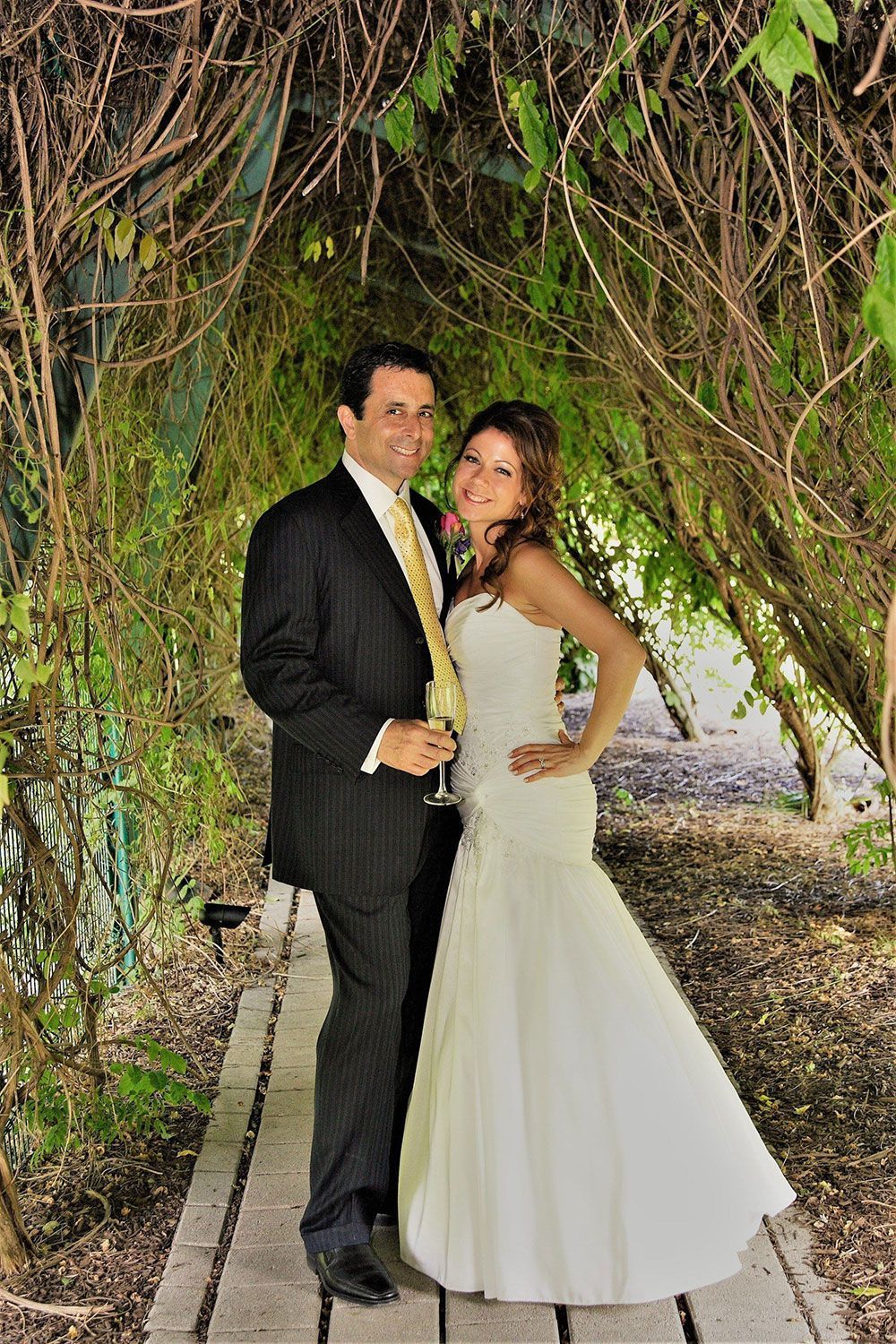 Wedding couple posing under a vine-covered arbor. The bride wears a strapless white gown, and the groom a suit.
