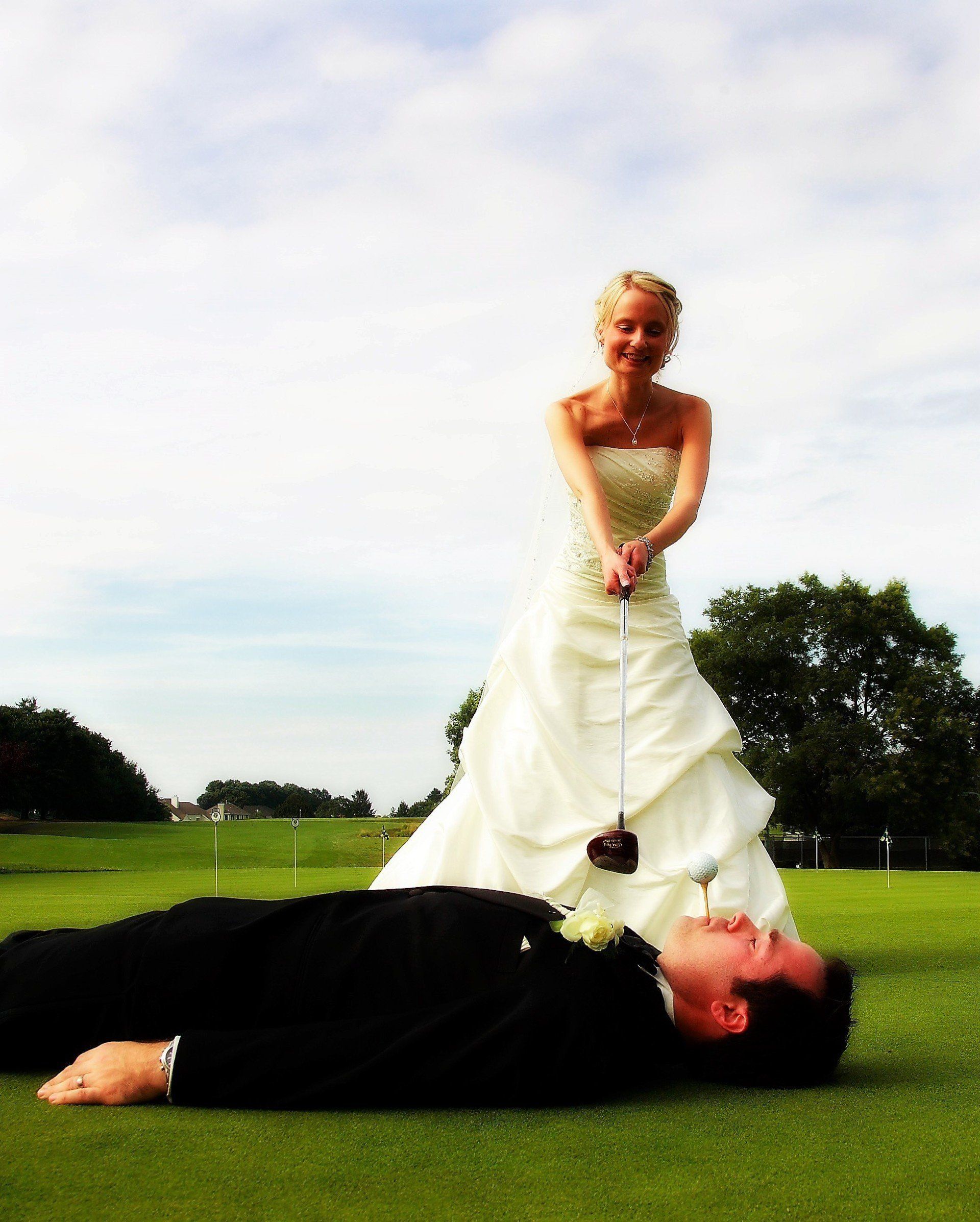 Bride in wedding dress, smiling, about to hit a golf ball with a club over groom lying on the grass.