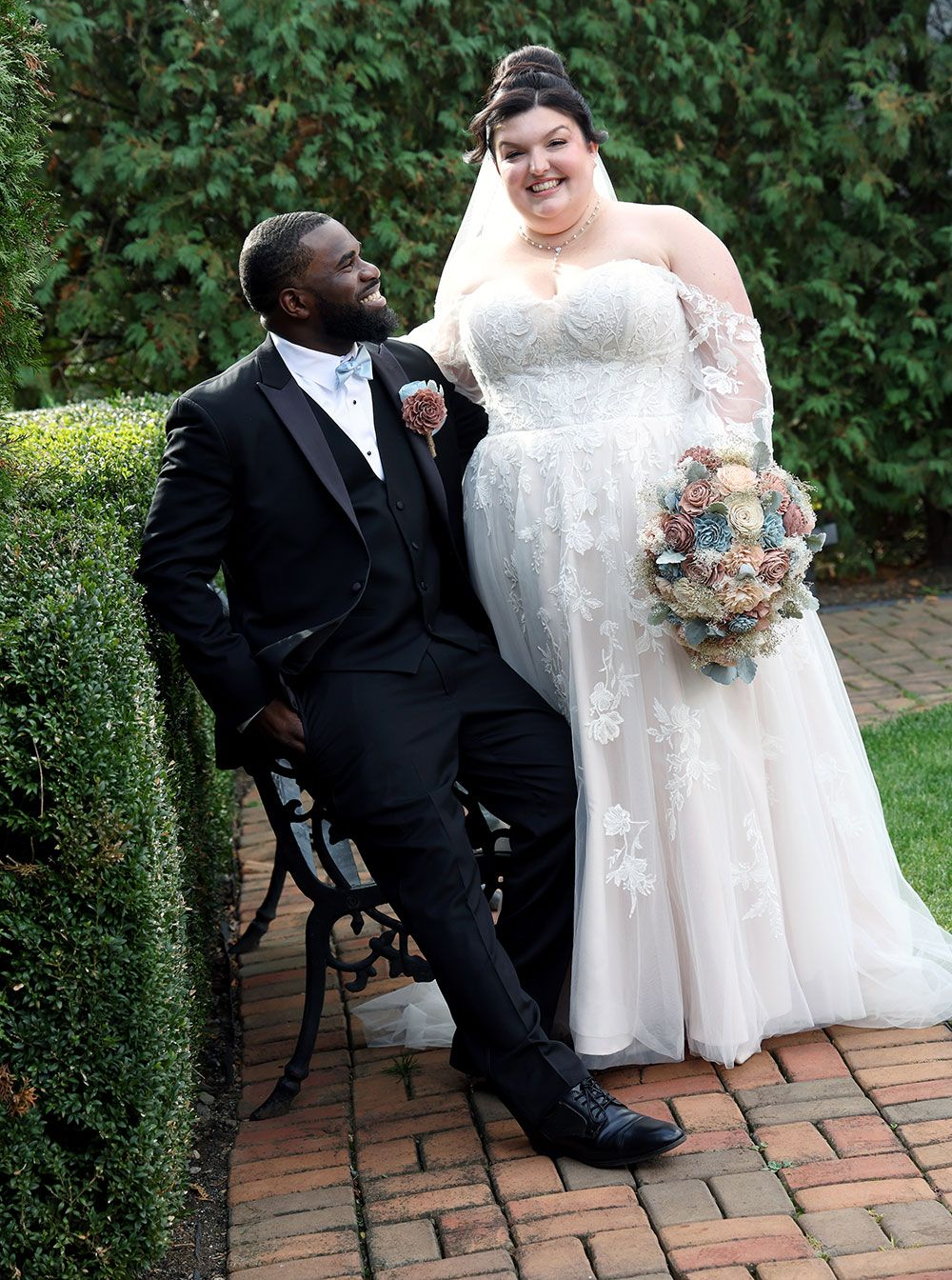Wedding couple smiling outdoors, groom in a suit and bride in a white gown holding bouquet.