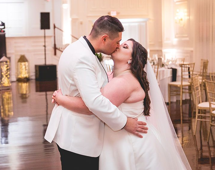 Couple kissing at a wedding reception; room with dance floor, tables, and decorations; soft lighting.