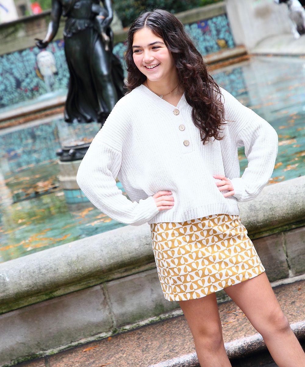 Woman in white sweater and yellow patterned skirt, posing in front of a fountain.