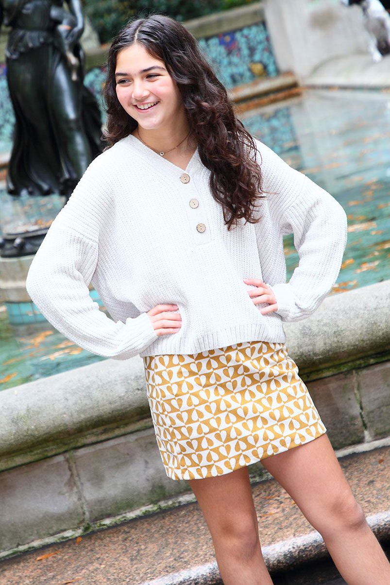 Woman in white sweater and yellow skirt poses by fountain.