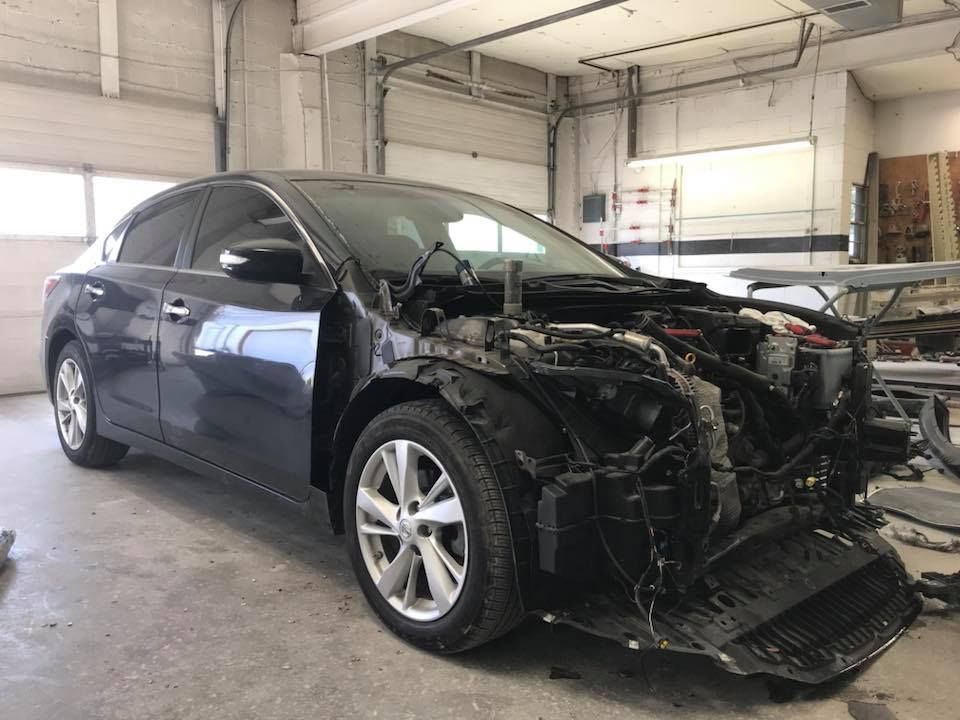 Dark gray sedan with front end damage in a repair shop.