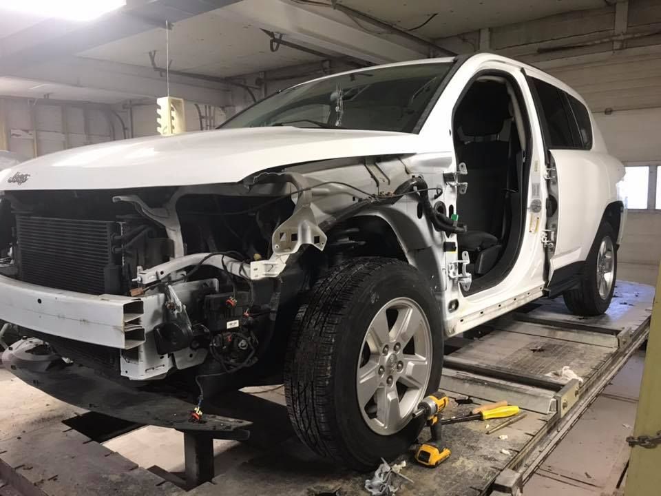 White SUV in a repair shop; front end damaged, parts removed.
