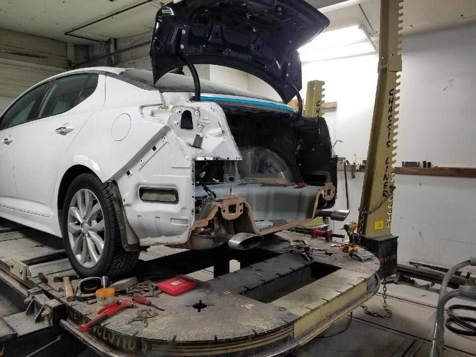 White car with rear damage in a repair shop, trunk open, surrounded by tools.