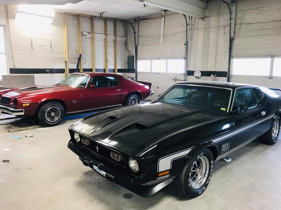 Black Ford Mustang Mach 1 and red Chevy Camaro muscle cars in a garage.