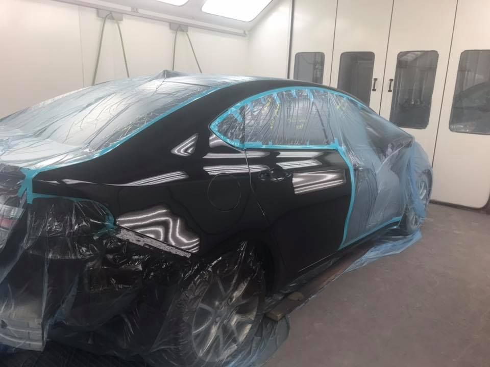Black car partially taped and covered in plastic, inside a paint booth.