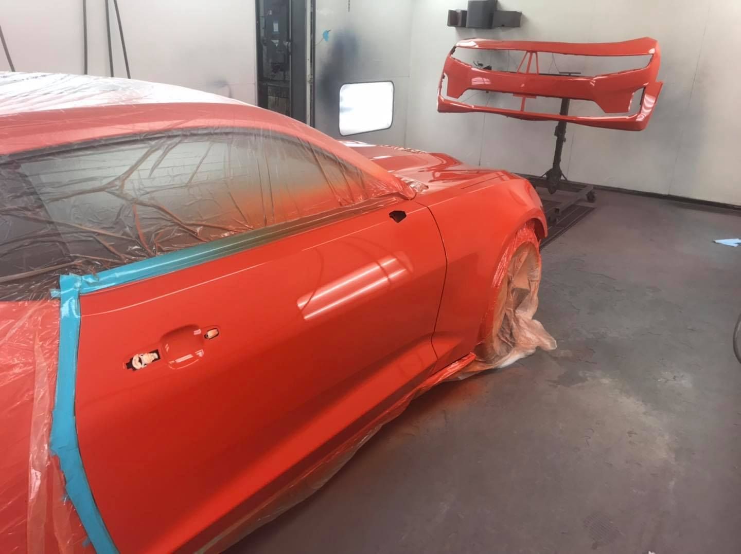 Red Camaro car being painted in a body shop; a detached bumper is in the background.