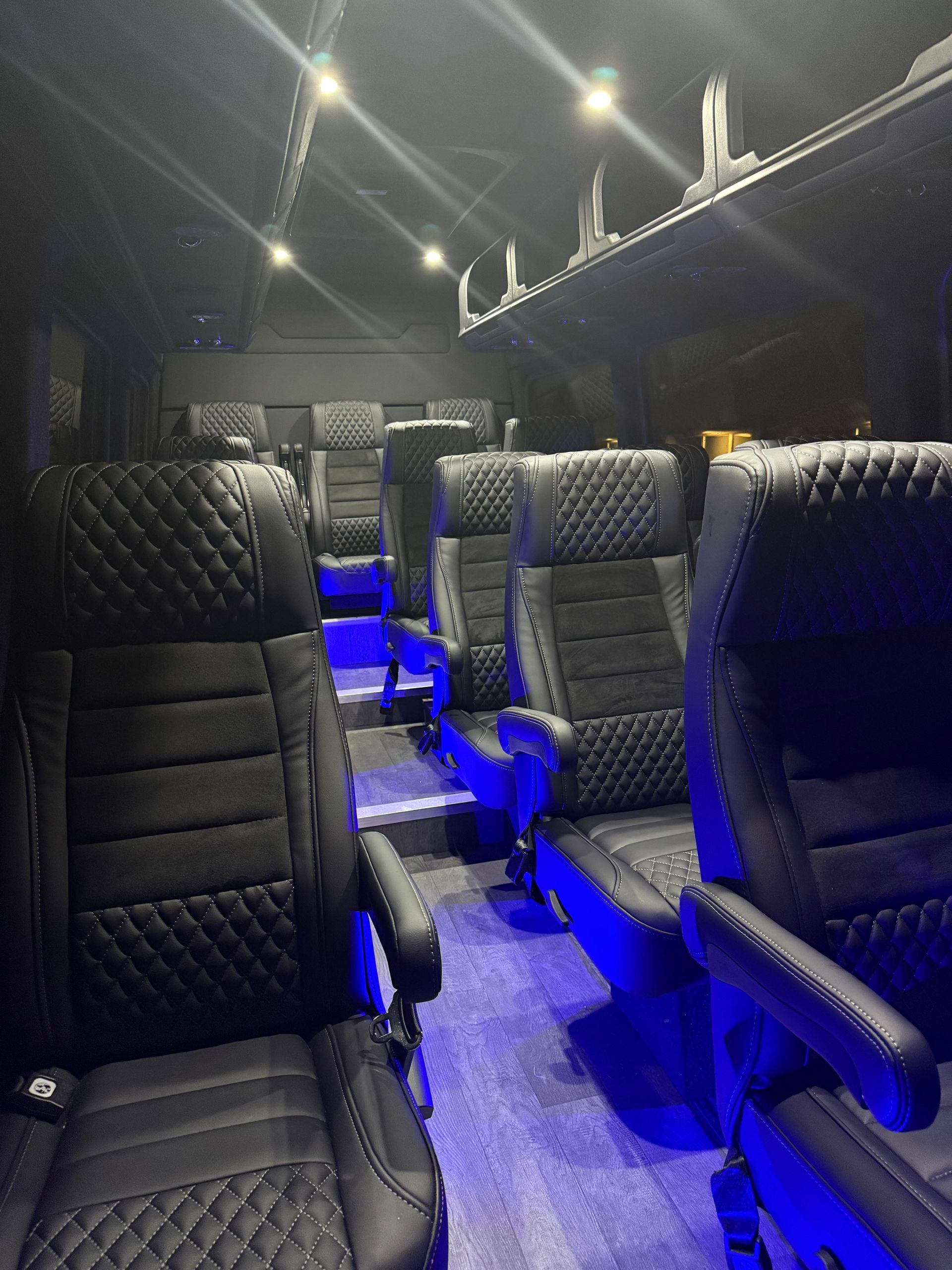 Interior of a luxury black passenger van with quilted leather seats, blue LED lighting, and overhead storage.