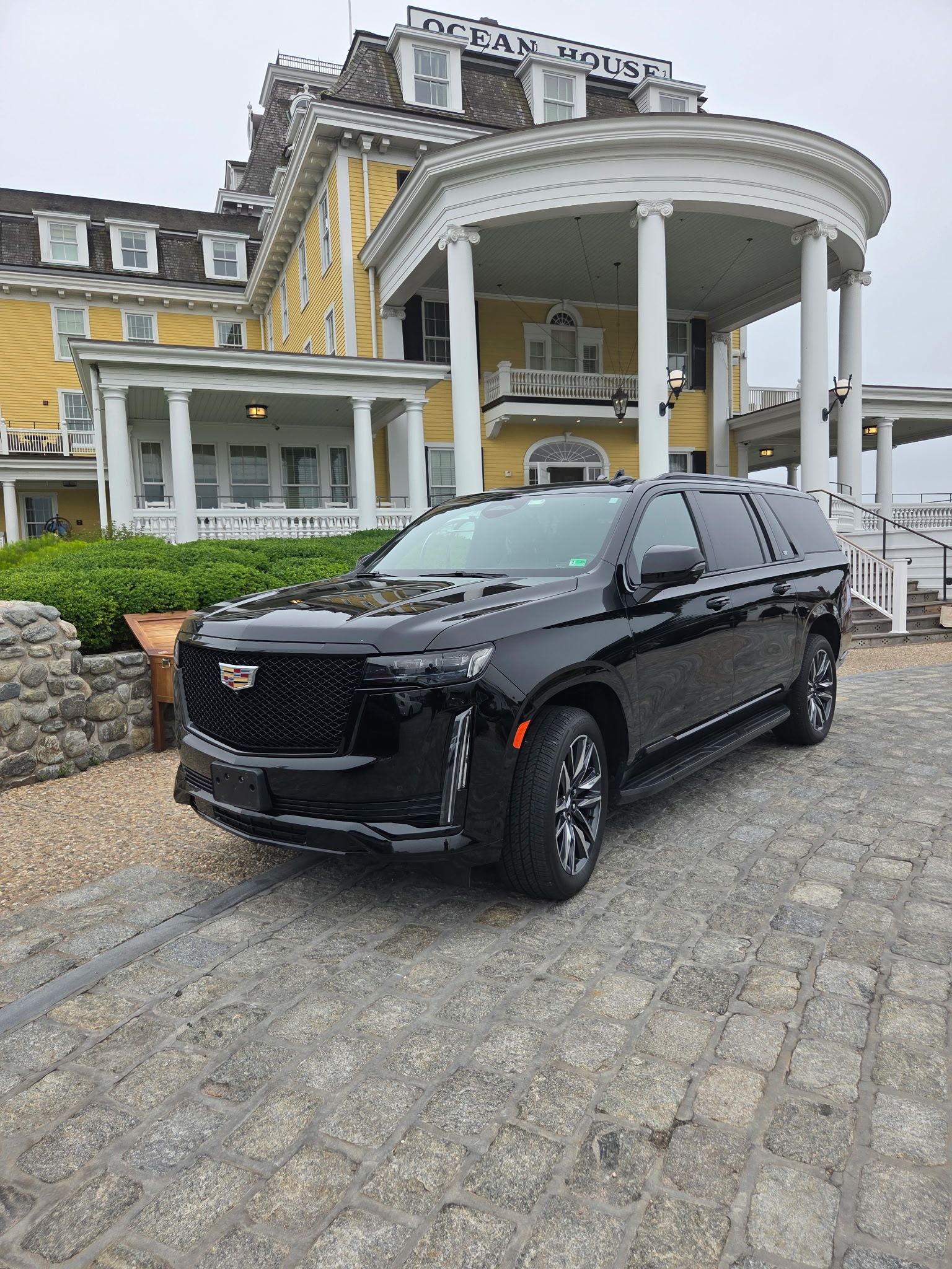 Black Cadillac SUV parked in front of a yellow colonial-style building with white columns and a stone wall.