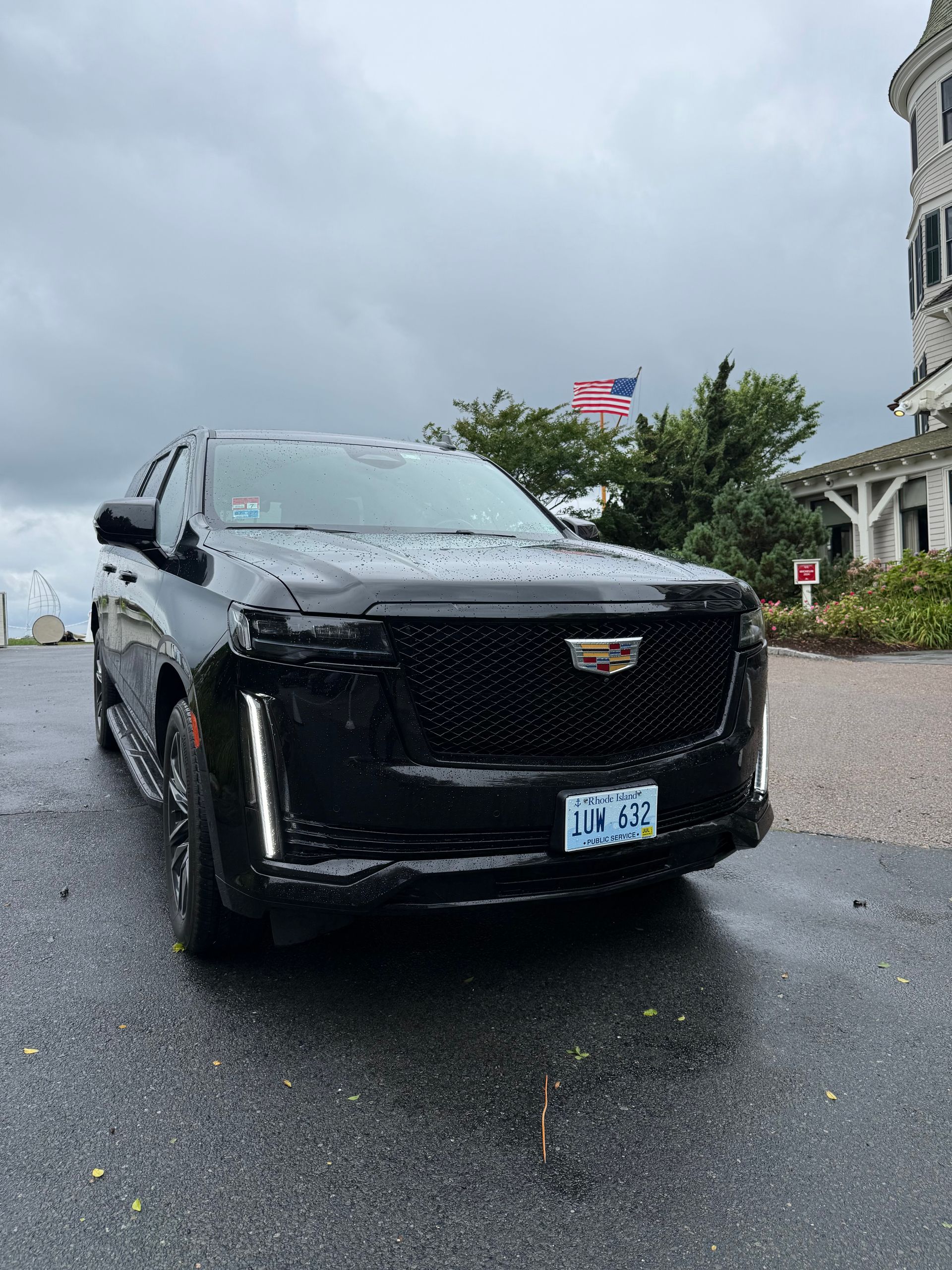 Black Cadillac Escalade parked near a building with an American flag on a cloudy day.