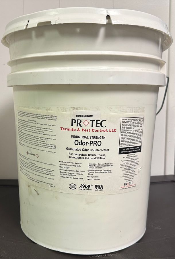 A white five-gallon bucket with a product label for Pro-Tec Odor-PRO, standing on a dark surface against a white wall.