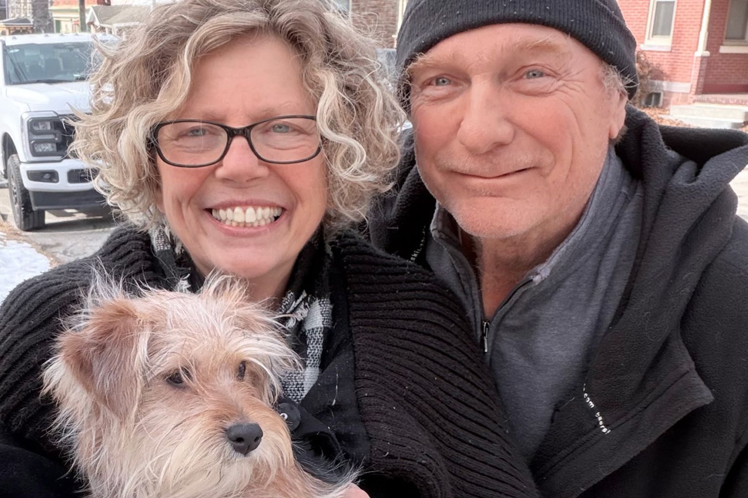 A smiling woman with curly hair and glasses holds a small, light-brown dog next to a smiling man in a black beanie.