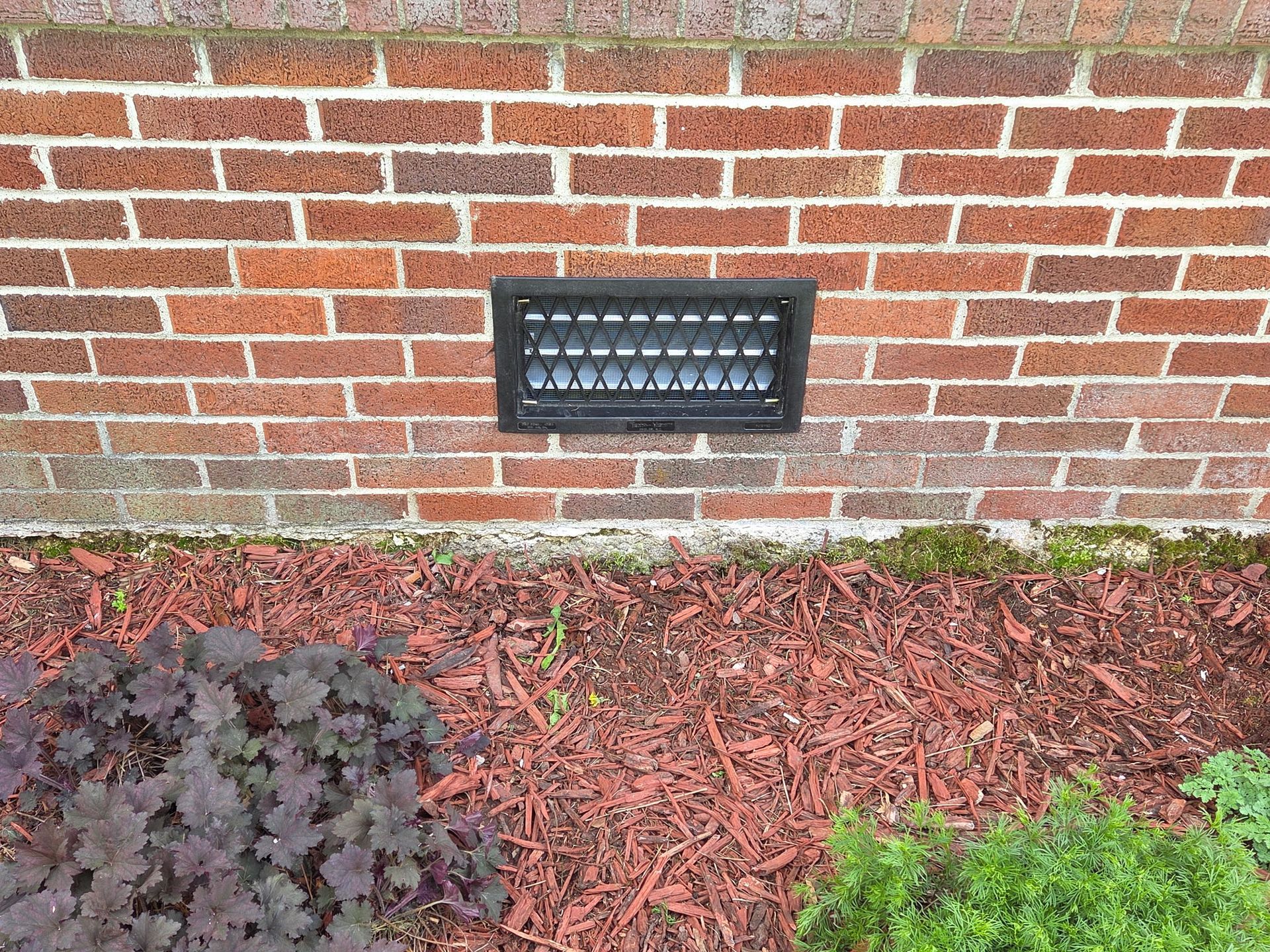Brick wall with a black outdoor vent and mulch bed below.