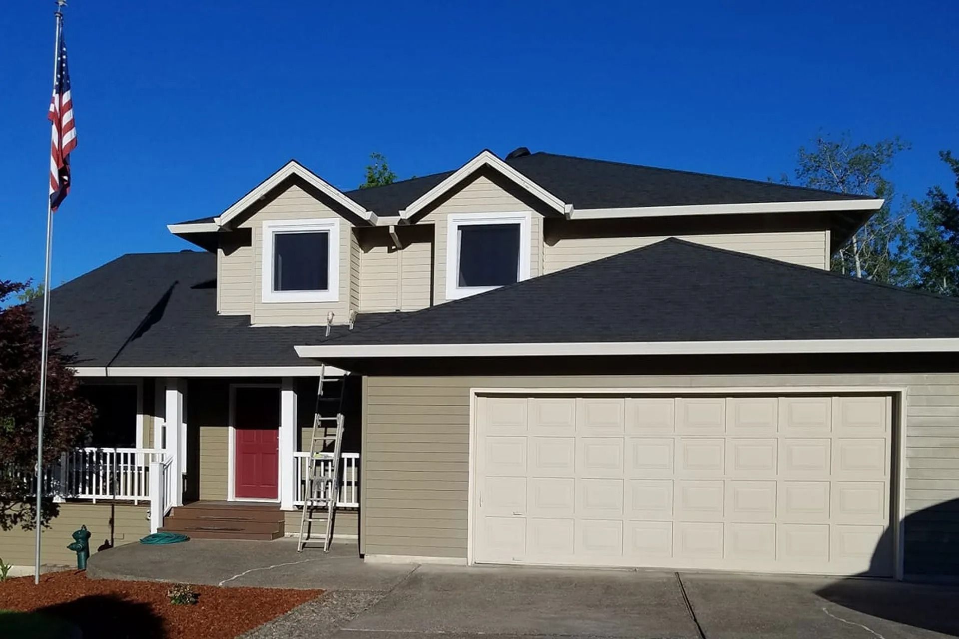 A two-story suburban house with tan siding, a dark roof, a red front door, and an American flag on a pole in the front yard.