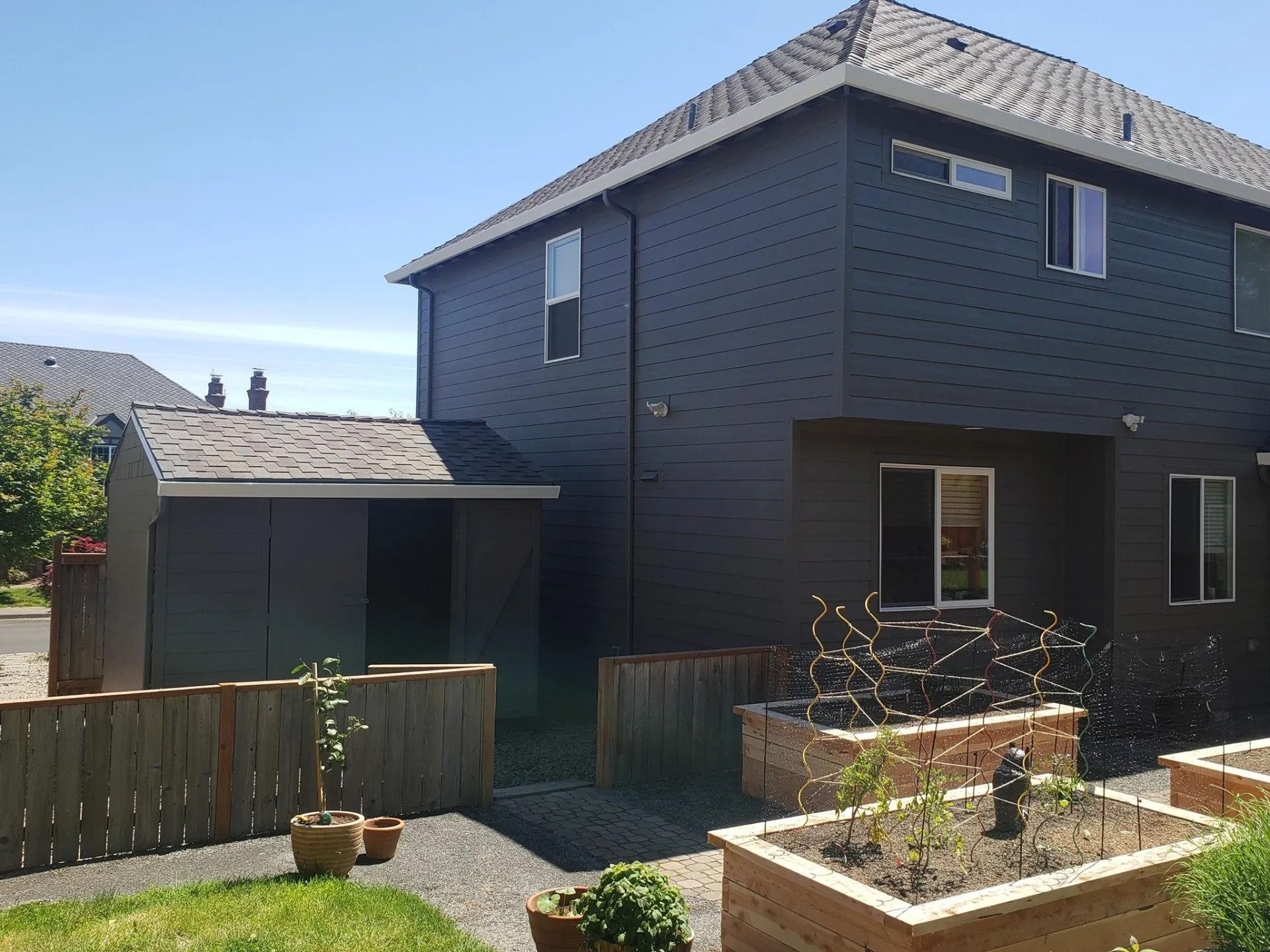 A side view of a dark gray house with a matching shed, wooden fence, and raised garden beds in the foreground.