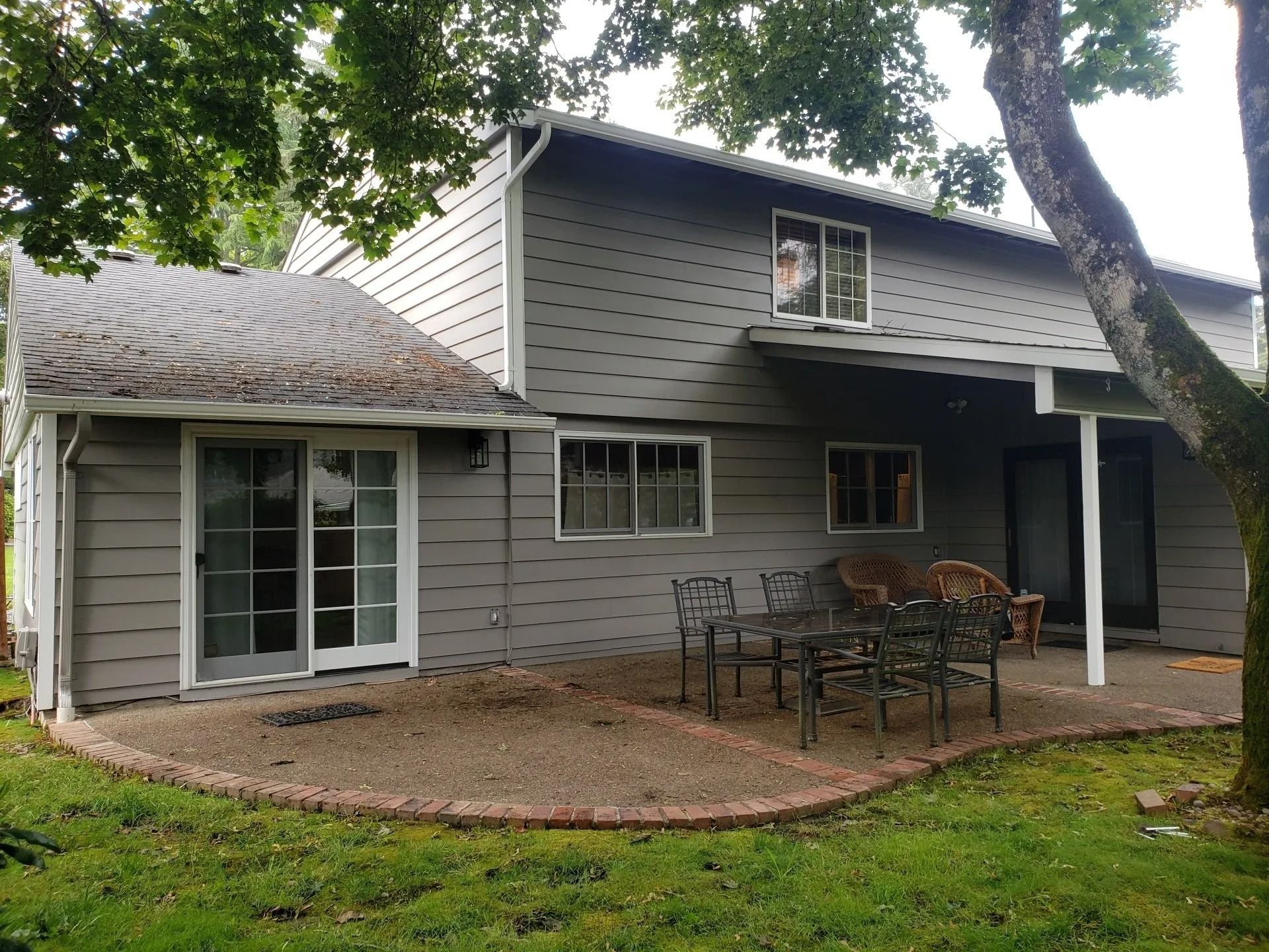 A grey two-story house with a backyard patio featuring a dining set, surrounded by a lawn and trees.