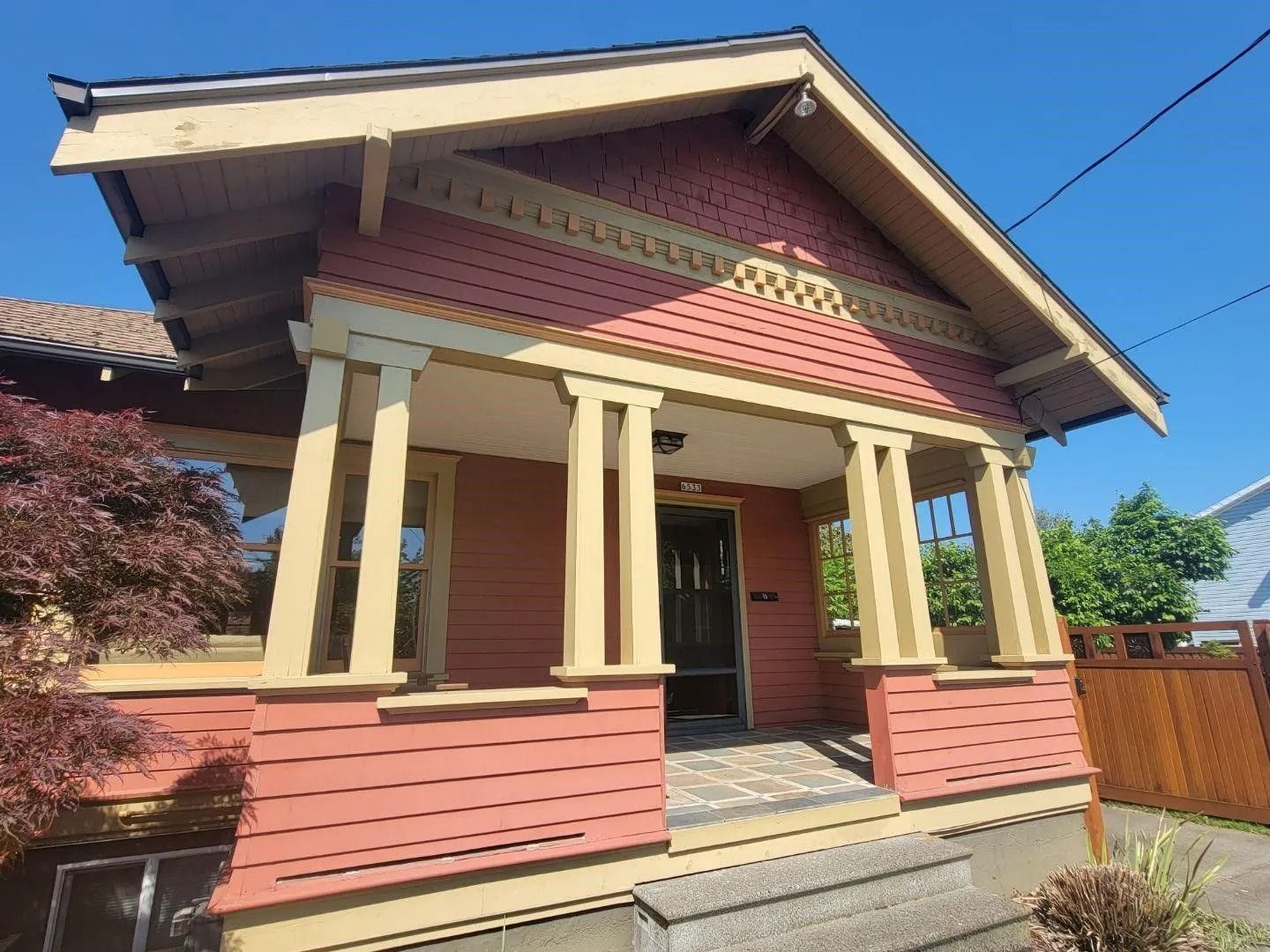 A small, red bungalow with cream-colored trim and a pillared front porch under a clear blue sky.