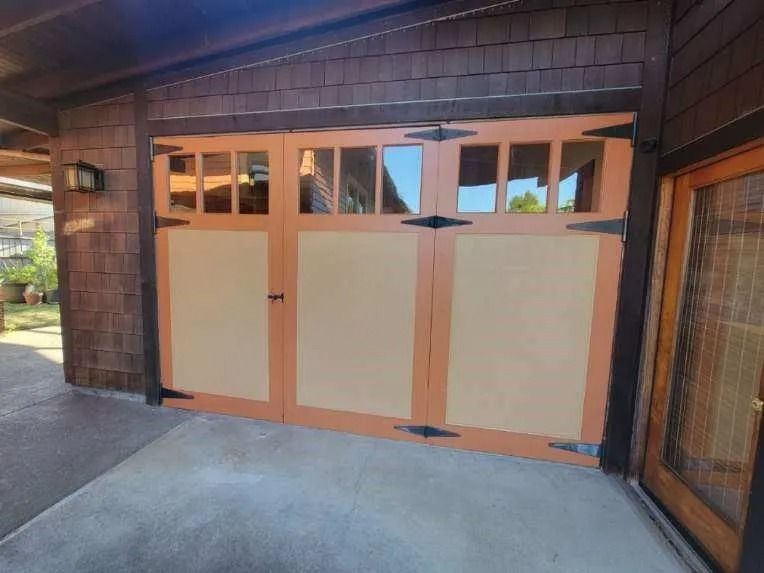 A tan-colored wooden garage door with windows and decorative black hinges, attached to a house with shingled siding.