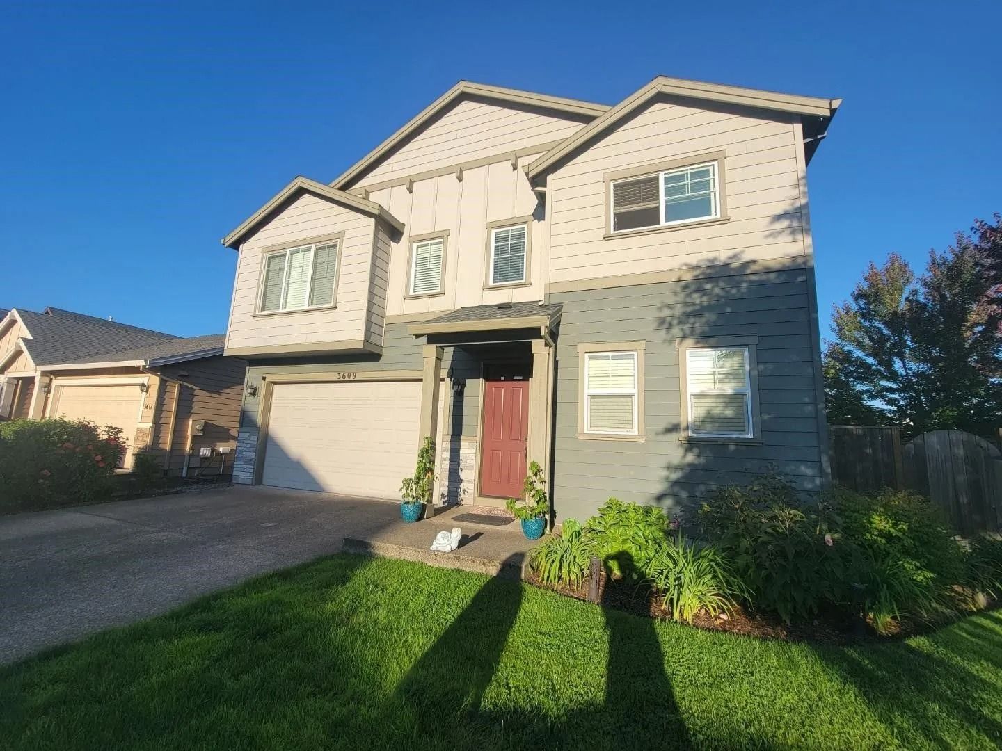 Two-story house with light beige siding, grey stone lower level, red front door, garage, and a sunny green front lawn.