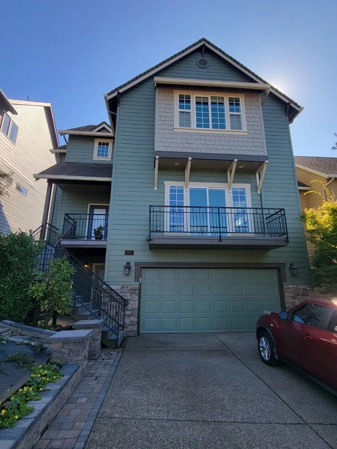 A three-story green house with a front-facing garage, a second-floor balcony, and a stone-accented foundation.