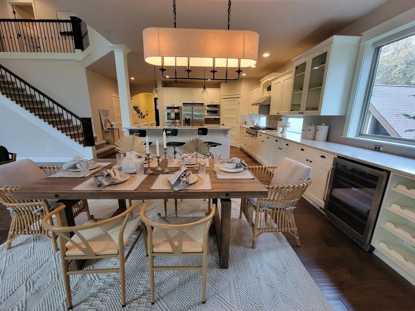 A modern, open-concept dining area and kitchen with a wooden table, woven chairs, and a large rectangular light fixture.