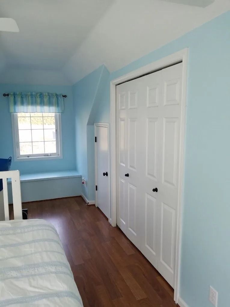 A blue-walled bedroom with a window seat, a white double-door closet, and wooden flooring next to a bed.